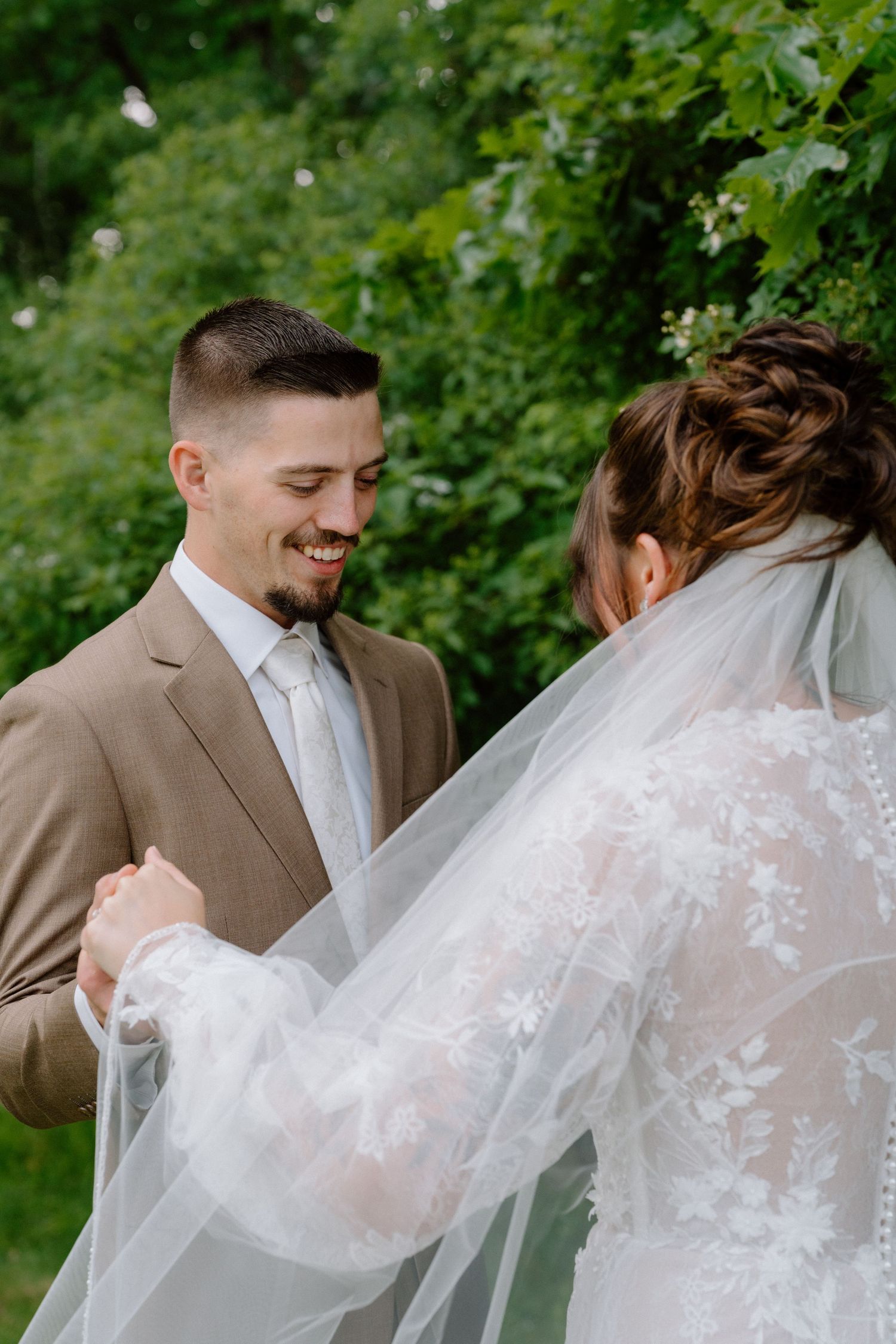Intimate wedding moment captured through lush greenery showing a couple in formal attire with delicate lace veil details.