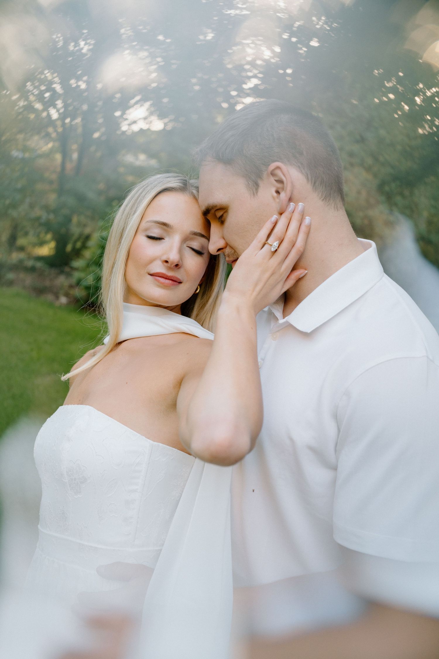 A romantic couple in white clothing embrace tenderly in a dreamy outdoor garden setting.