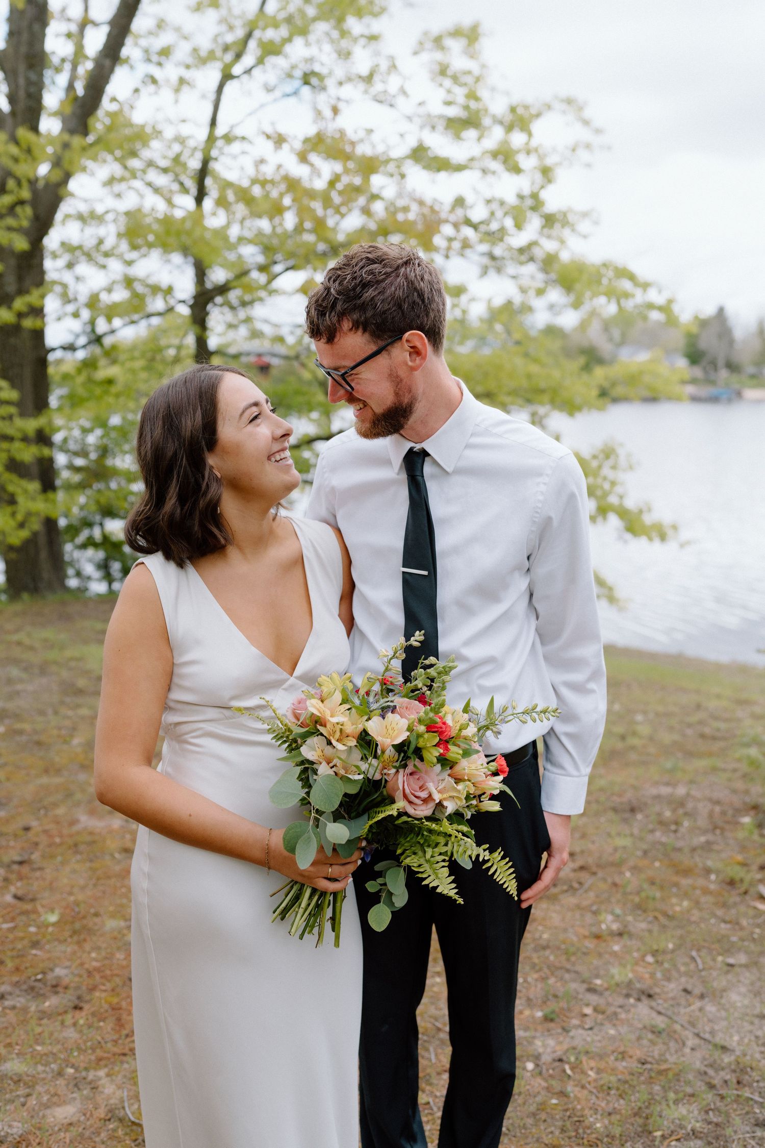 Newlyweds share intimate moments while holding a floral bouquet near a lakeside.