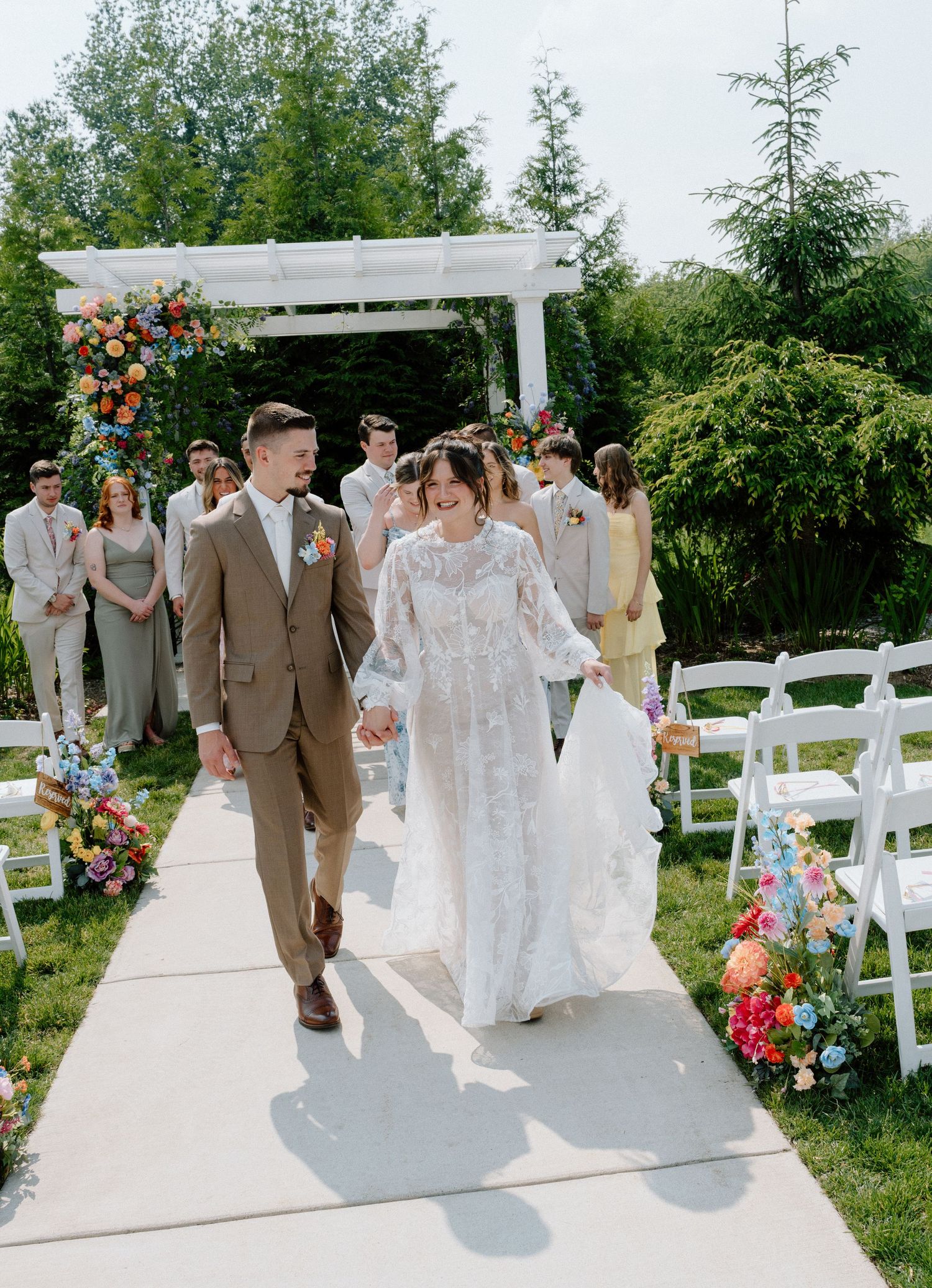 Wedding party walks down an outdoor aisle decorated with white chairs and floral arrangements.