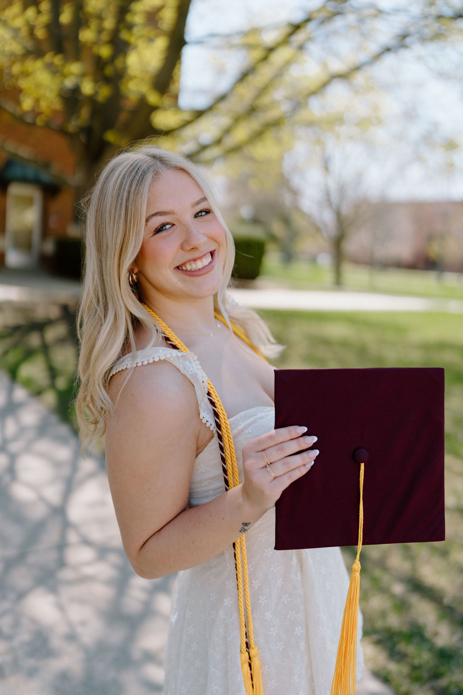 Graduate in white dress poses outdoors with gold honor cords and diploma.