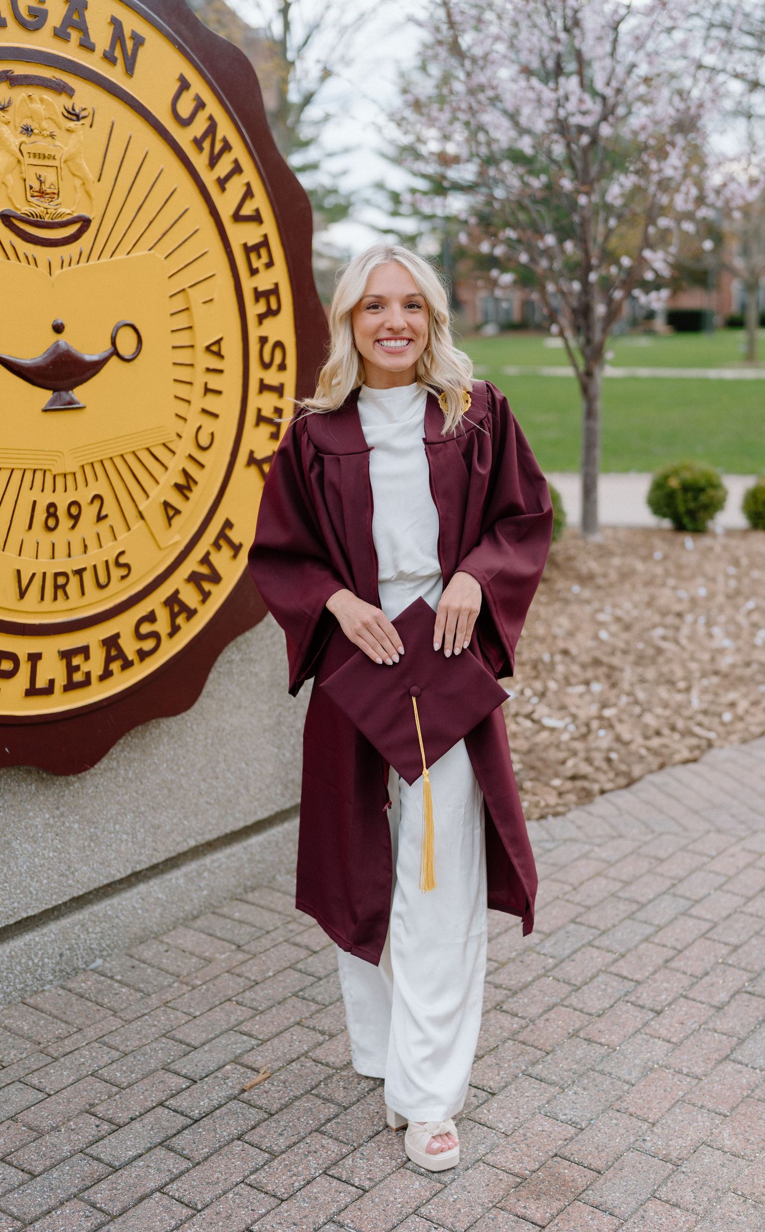 Graduate in burgundy cap and gown poses by university seal on campus.
