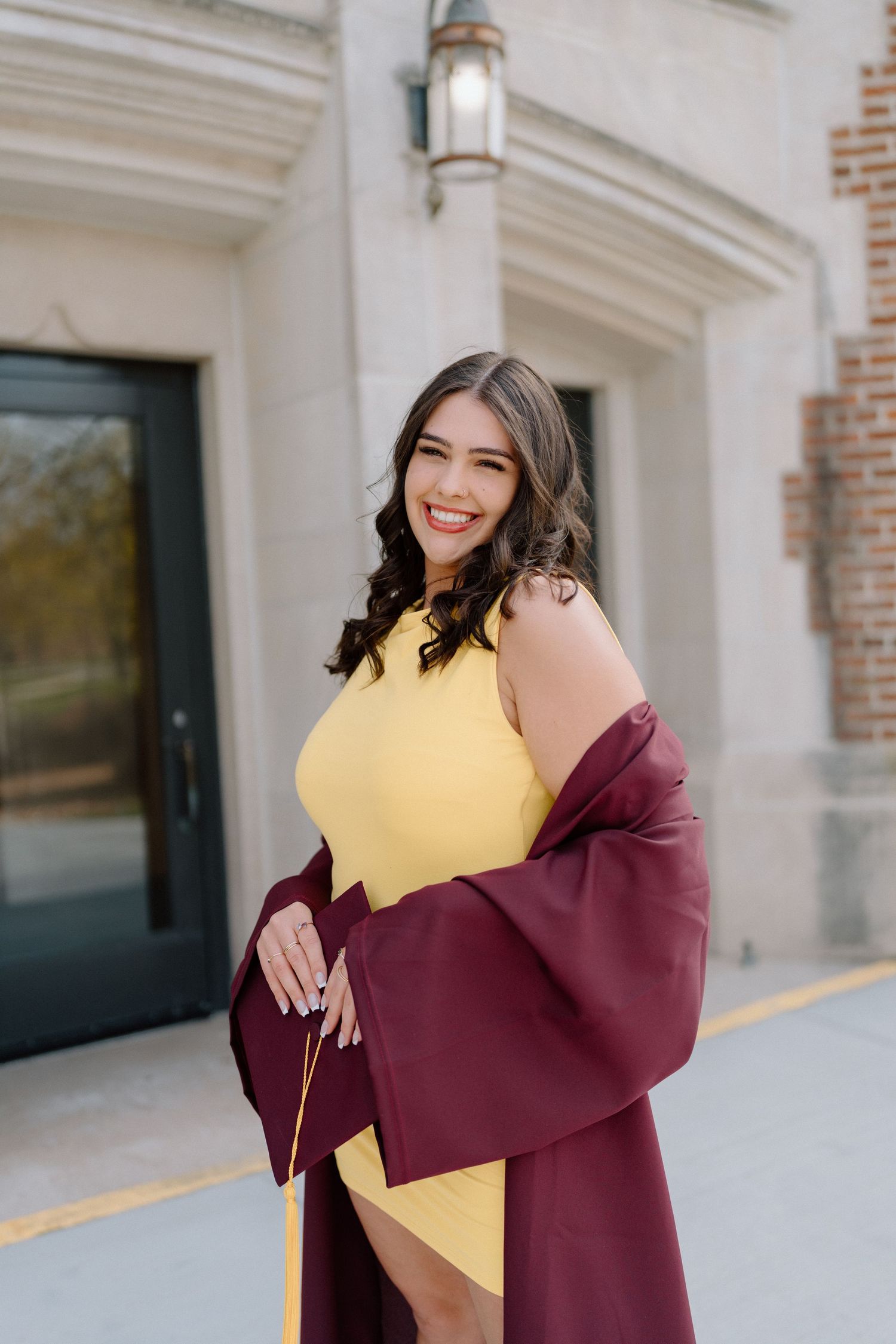 A joyful embrace between a graduate in burgundy gown and a family member in yellow against a white building exterior.