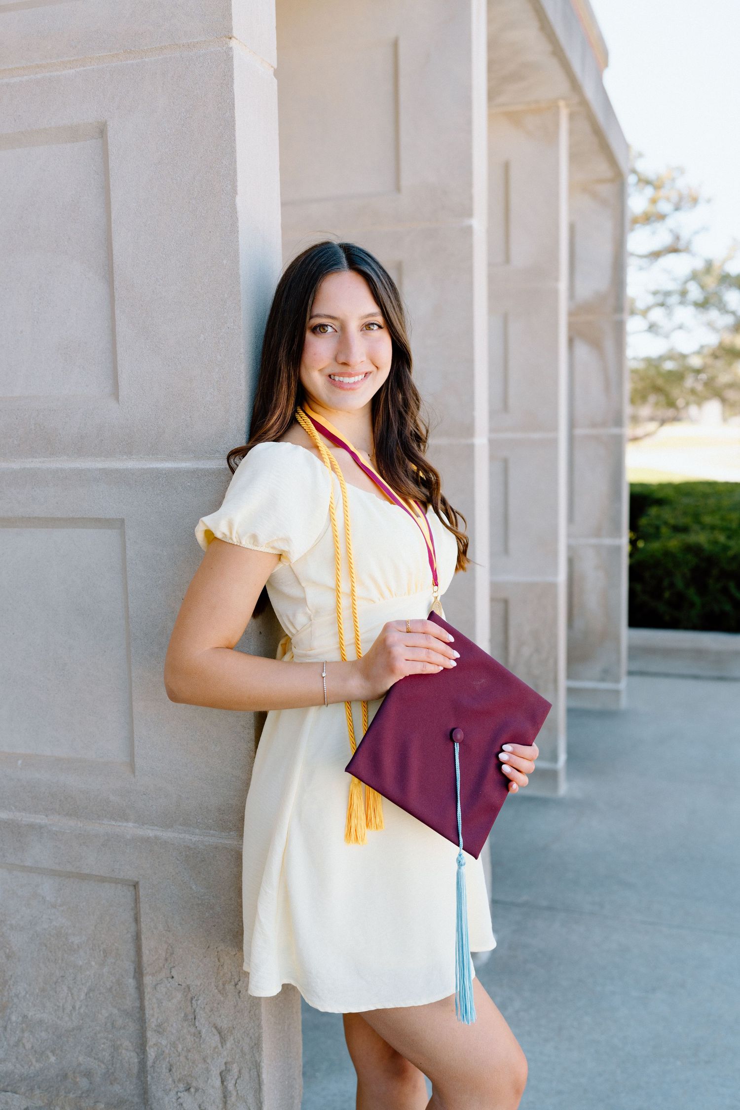 Smiling young woman in white dress holding maroon graduation cap with tassels, leaning against stone column outdoors.