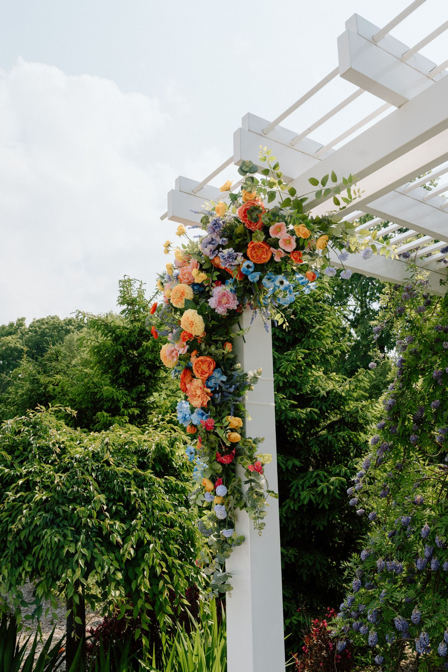 White pergola adorned with cascading coral and peach floral arrangements set against lush garden greenery.