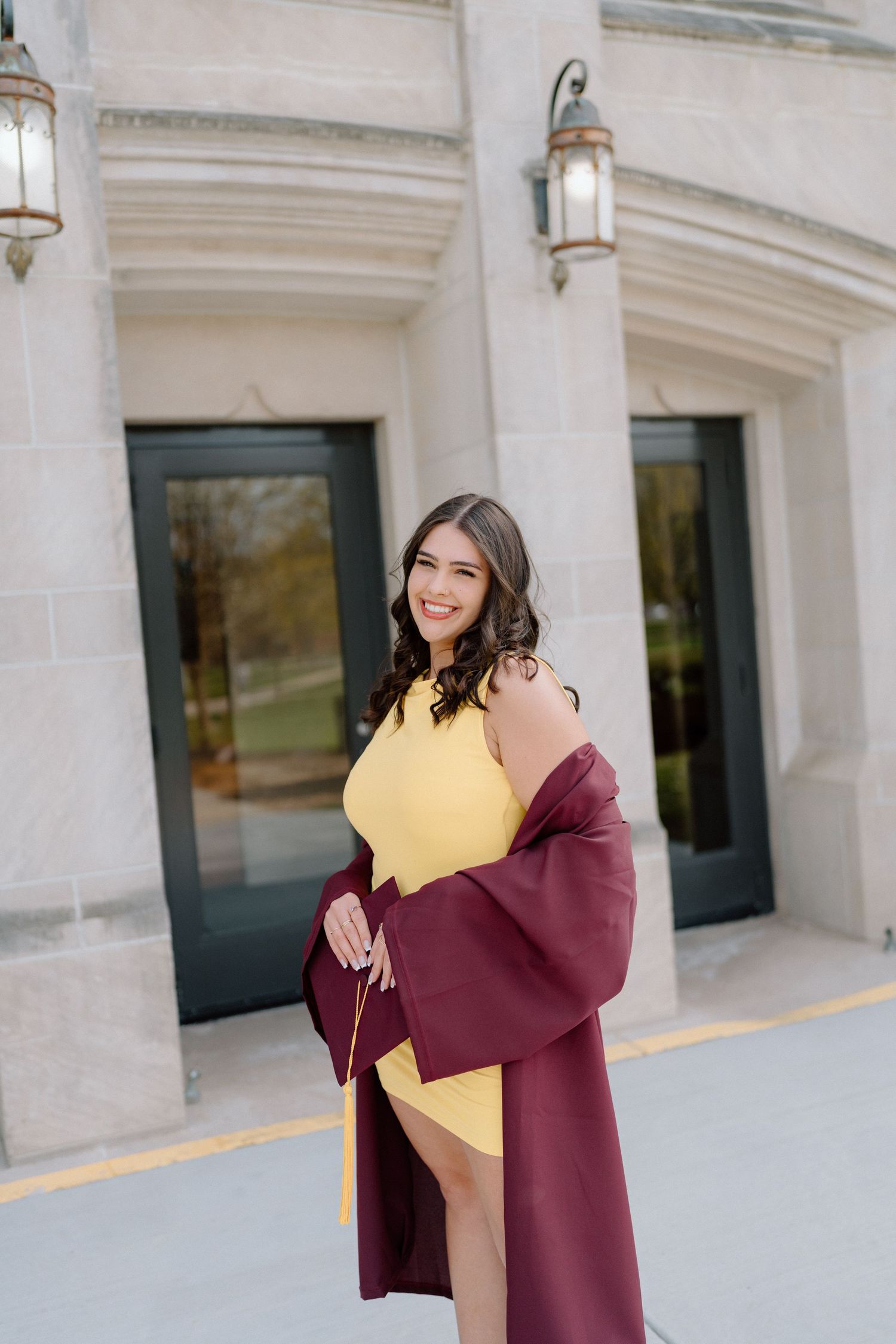 Graduate in maroon academic regalia poses against white stone colonnade architecture.