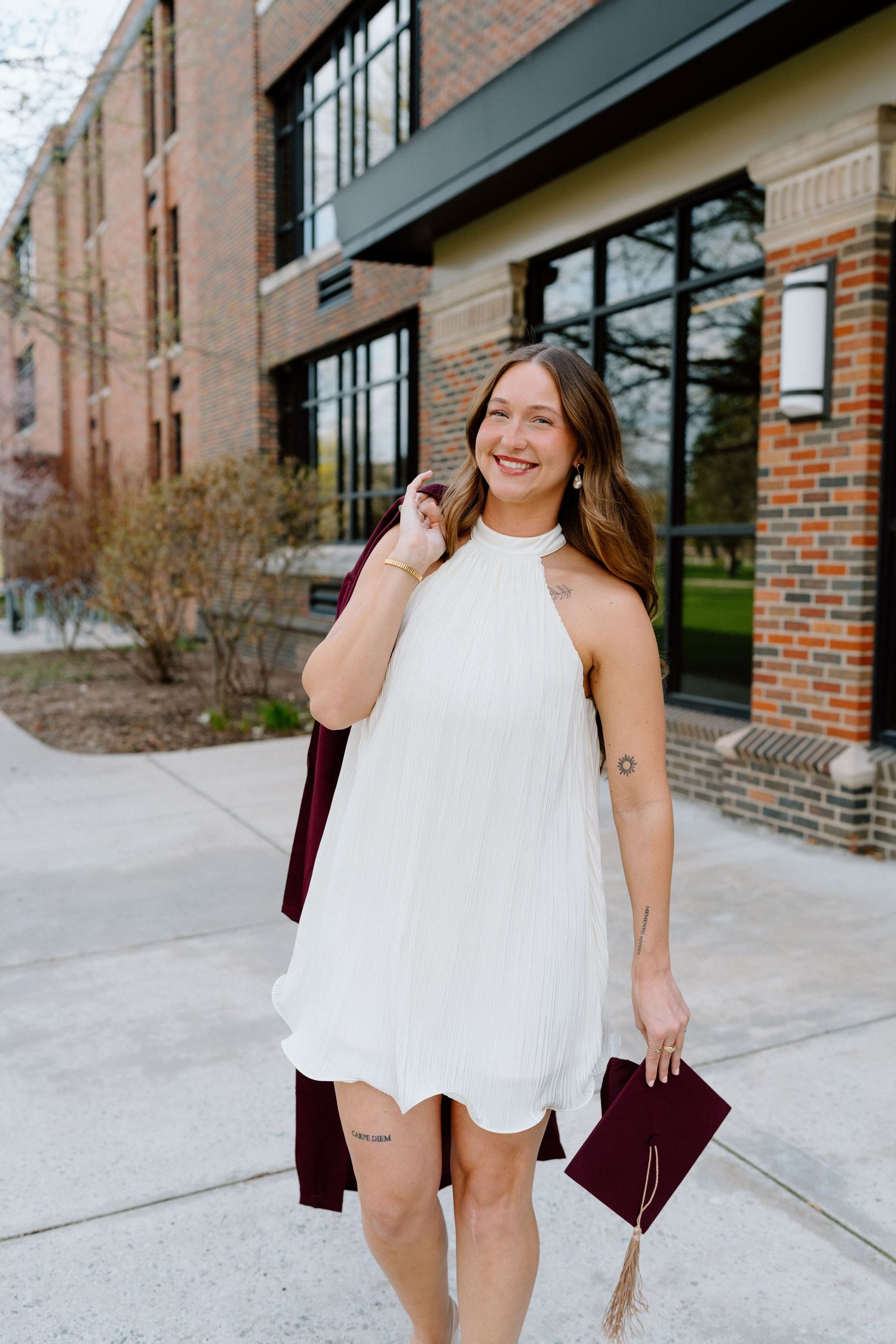 Smiling graduate in white halter dress holding cap and maroon gown outside brick university building.