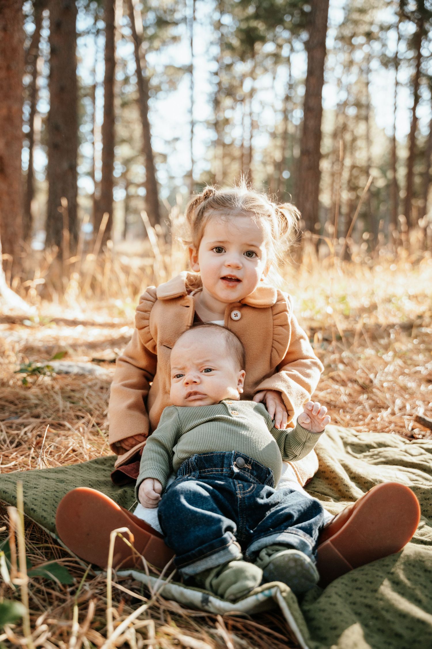Little child sitting with younger baby sibling on blanket in the woods.