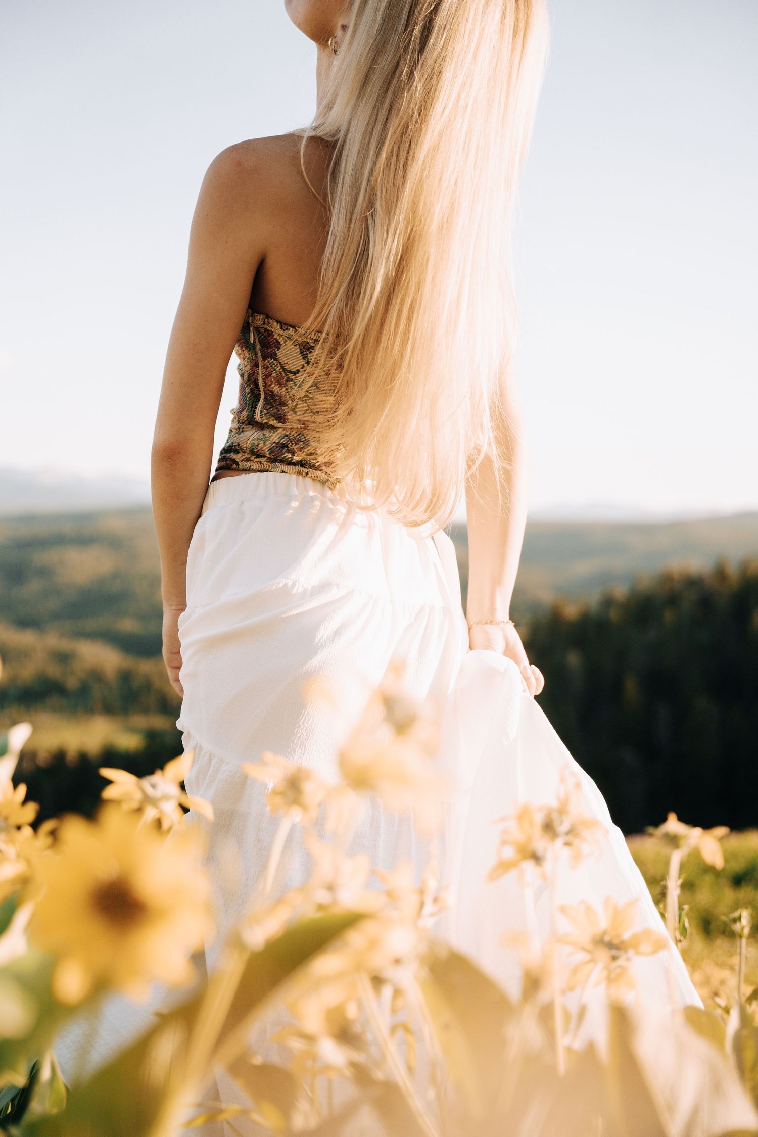 Girl propping her head up in the middle of a yellow flower field. Mountains and open green spaces in the background.