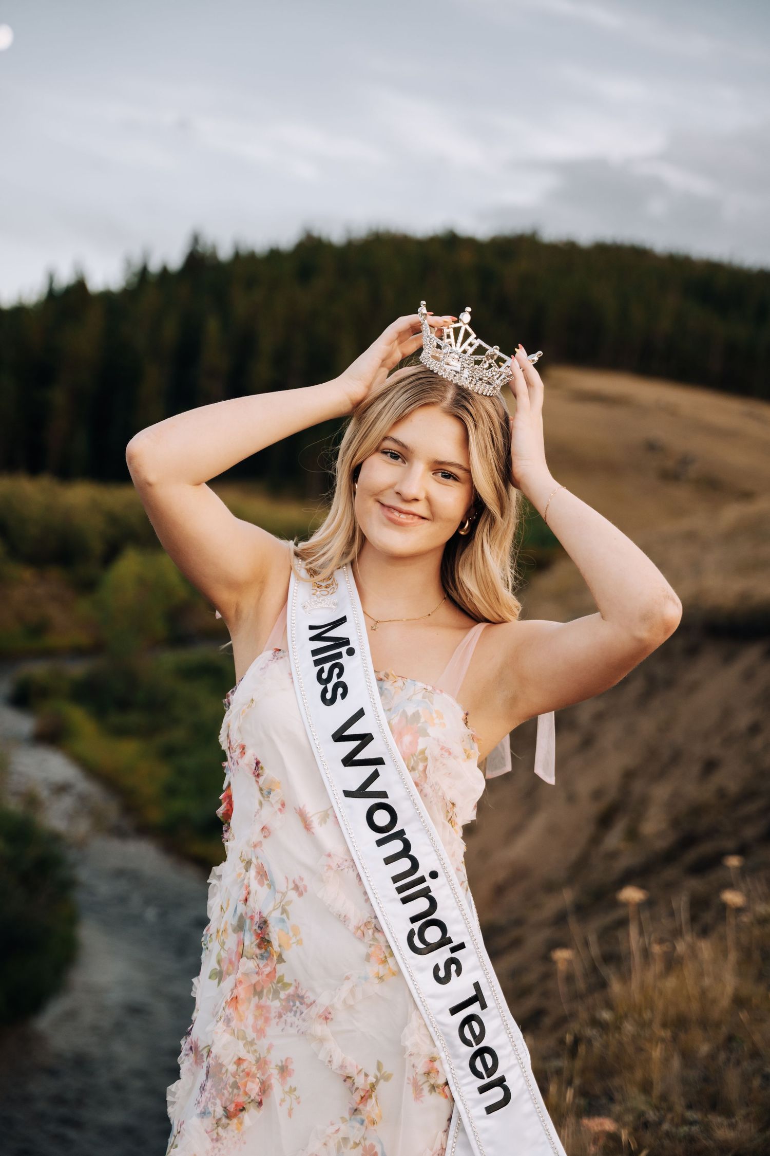 Girl holding a crown on top of her head while wearing a sash across her body.
