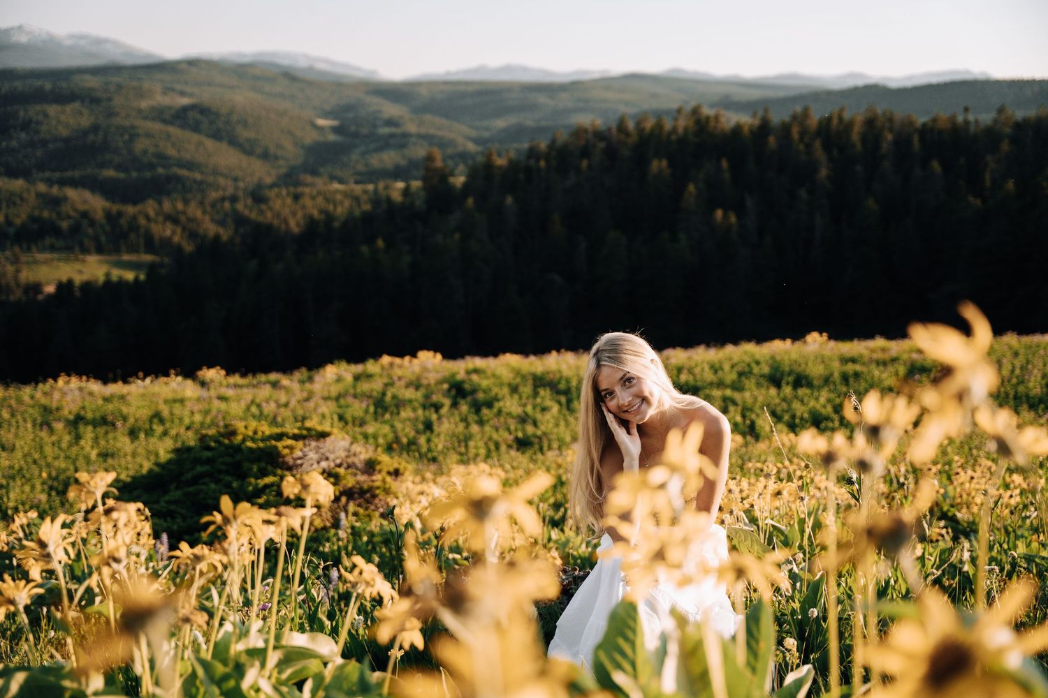 Girl propping her head up in the middle of a yellow flower field. Mountains and open green spaces in the background.