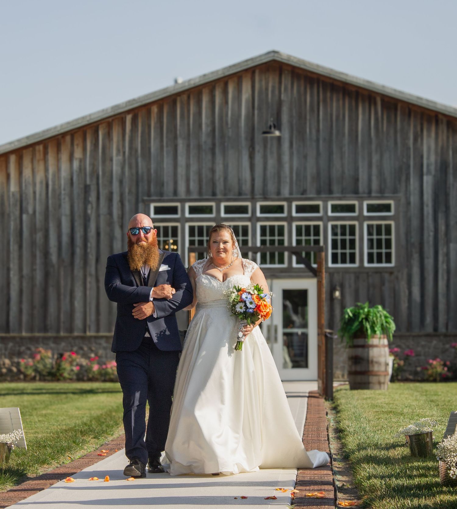 Bride and escort walk down pathway in front of rustic wooden barn venue.