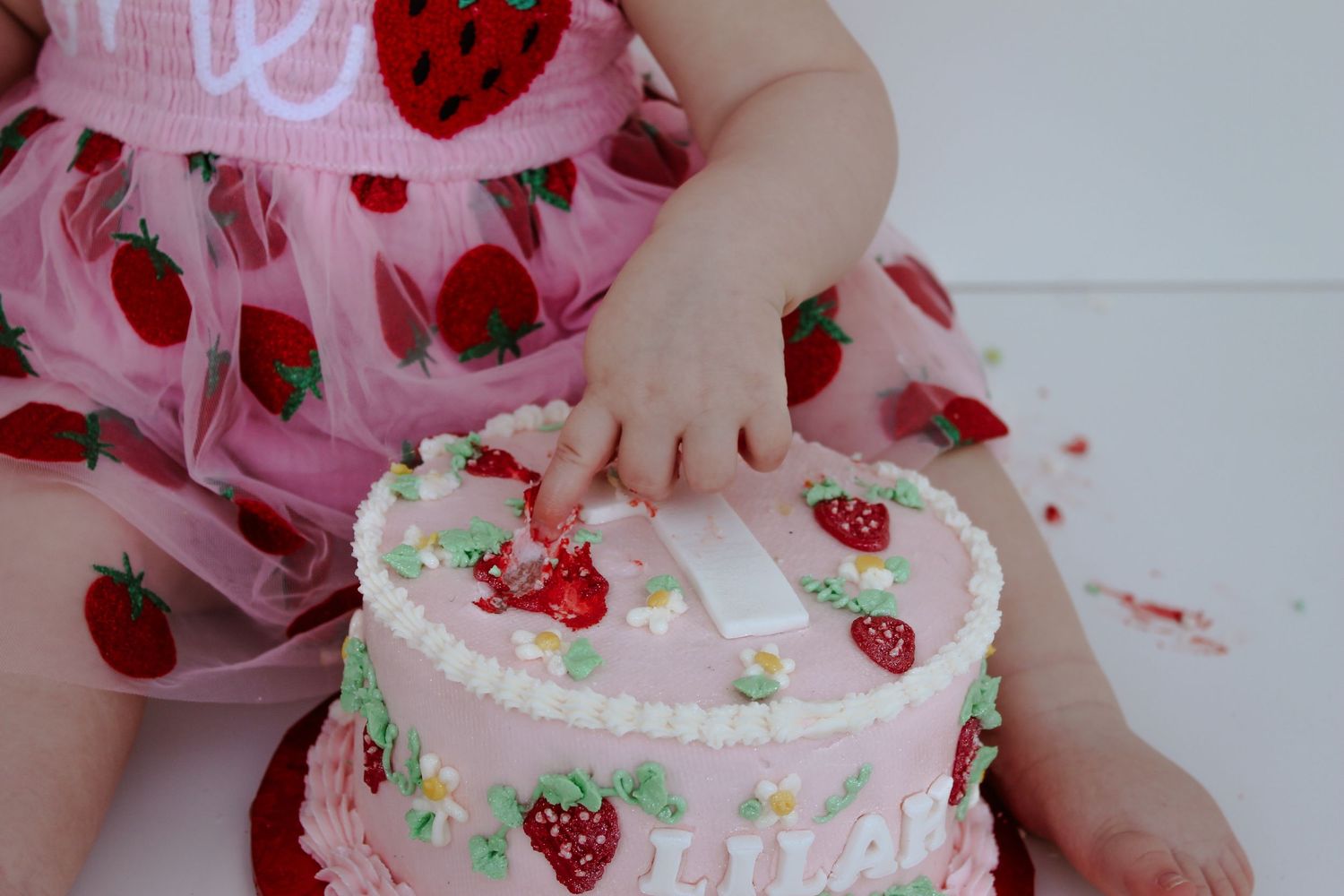 Pink birthday cake with strawberry decorations and small hands reaching for it.