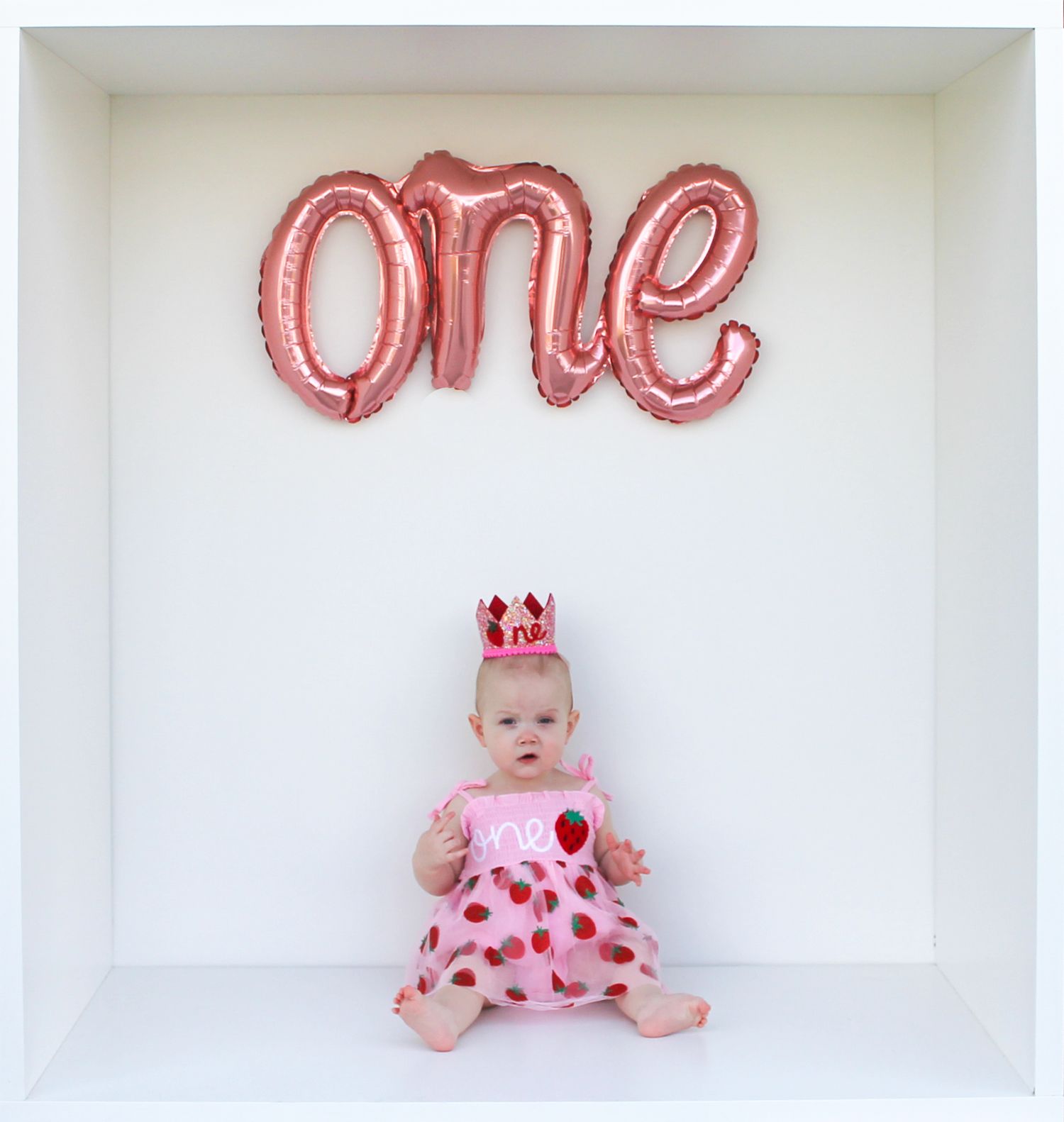 A baby wearing a pink polka dot dress and crown sits against a white backdrop beneath a red balloon 'one' text.