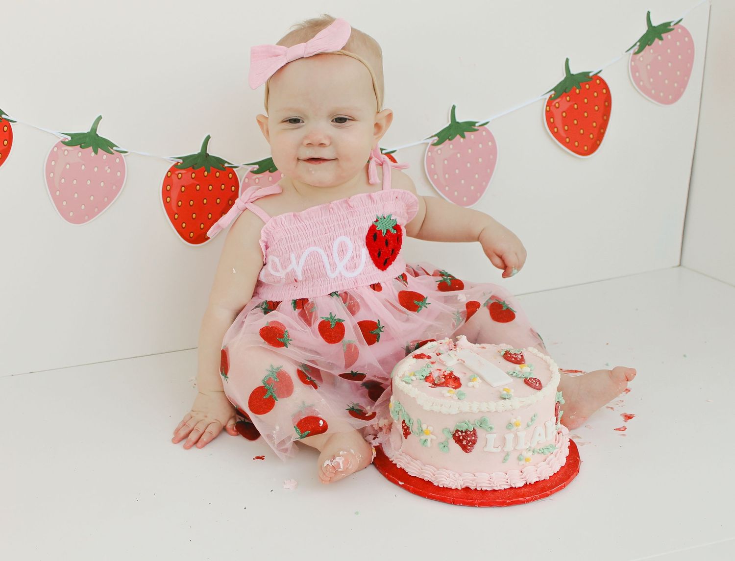 Baby in pink strawberry outfit reaching for decorative birthday cake on white background.