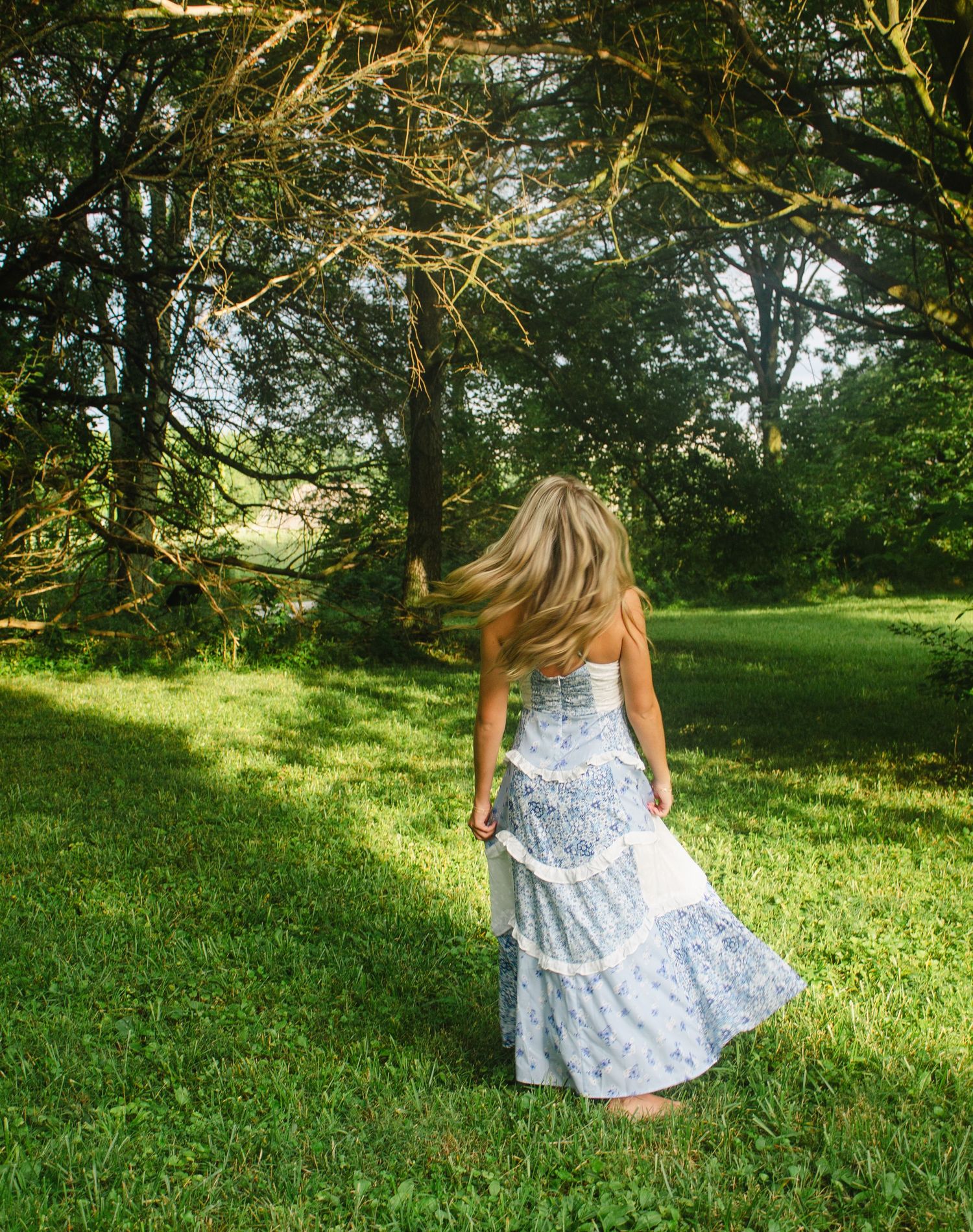 A person in a flowy white summer dress twirls in a sunlit garden with trees and lush grass in the background.