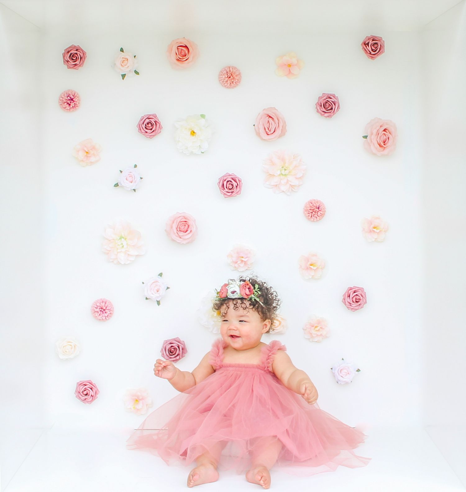 An infant in a pink tulle dress lies on white backdrop with soft pink floral wall decorations.