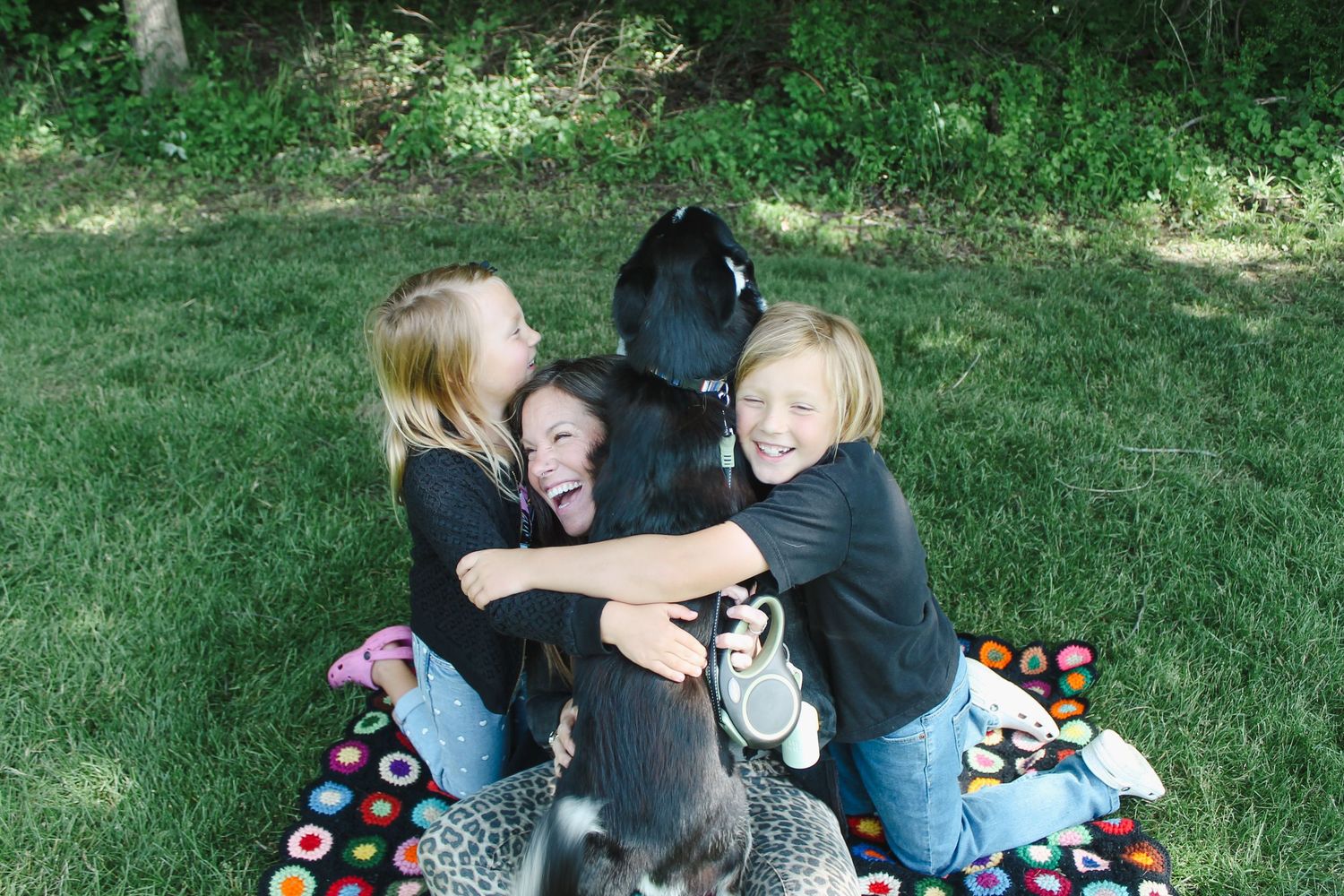 A black dog sits on a colorful blanket in the grass while three people hug it from all sides.