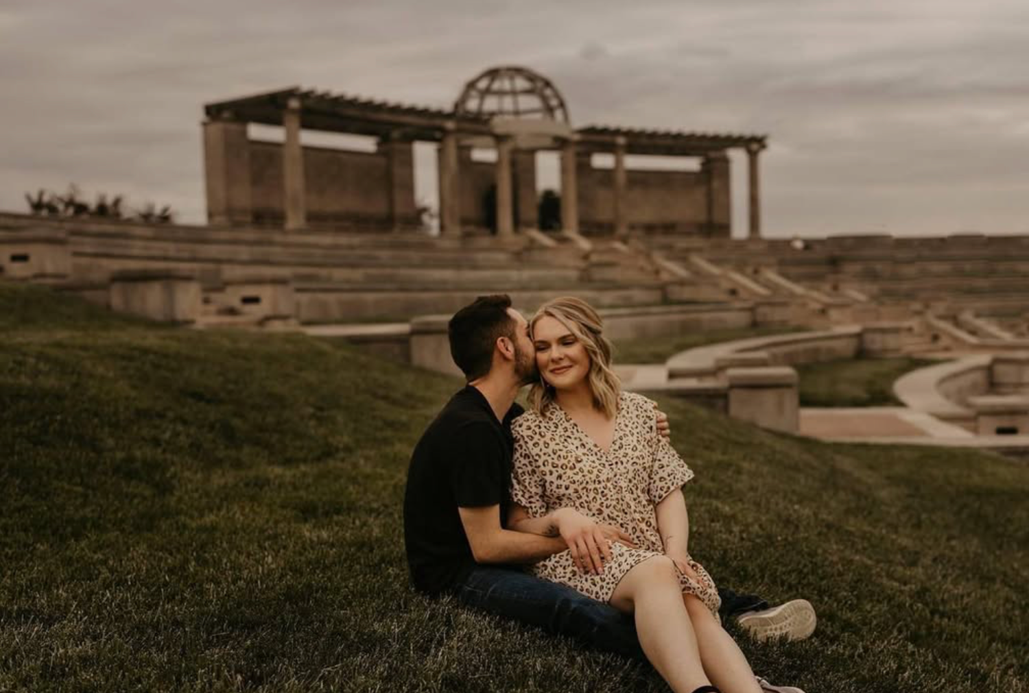 Couple sitting on grassy hill during golden hour with buildings in background.