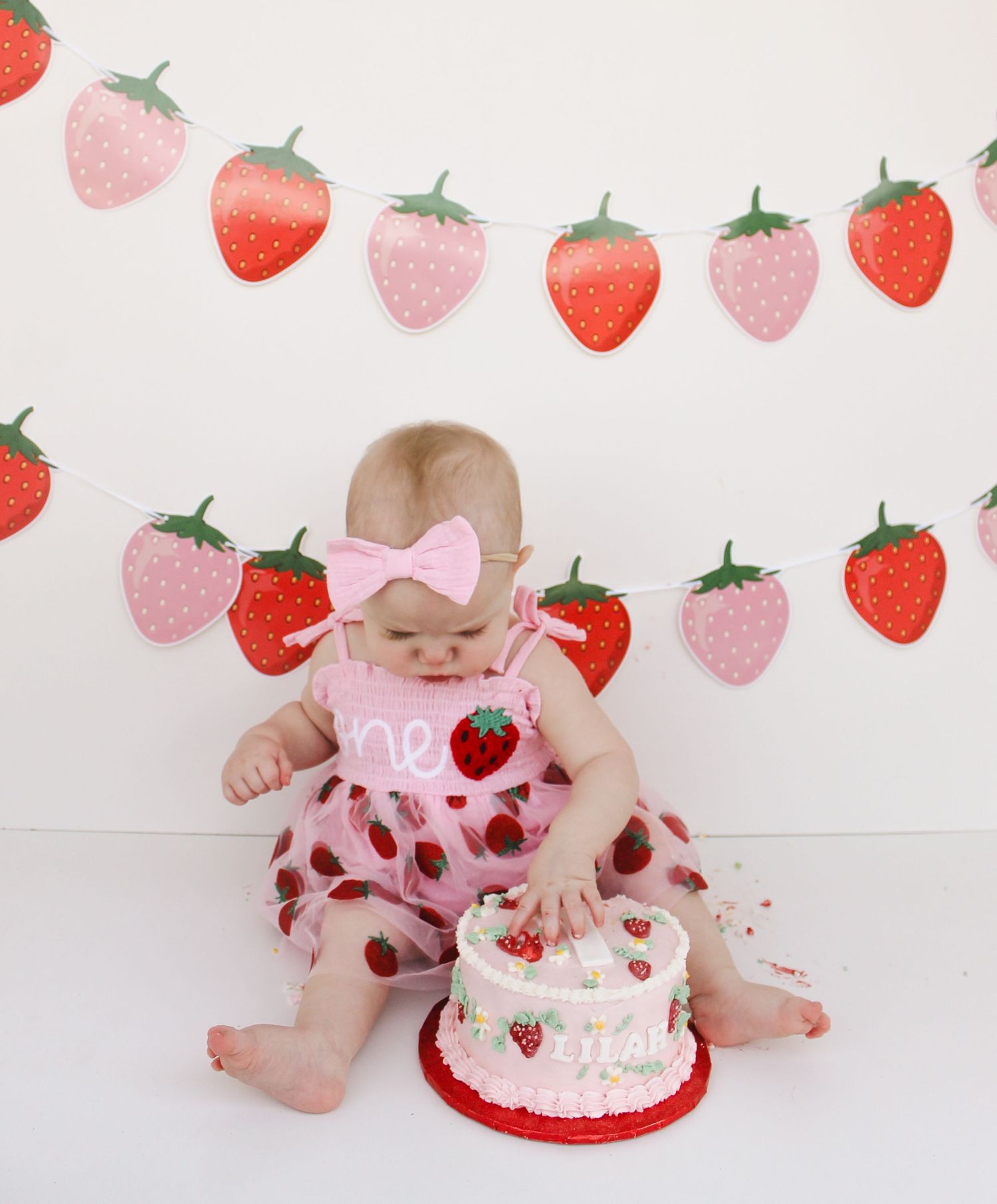 Baby in pink onesie with strawberry cake smash photo sequence against white backdrop with strawberry pattern.