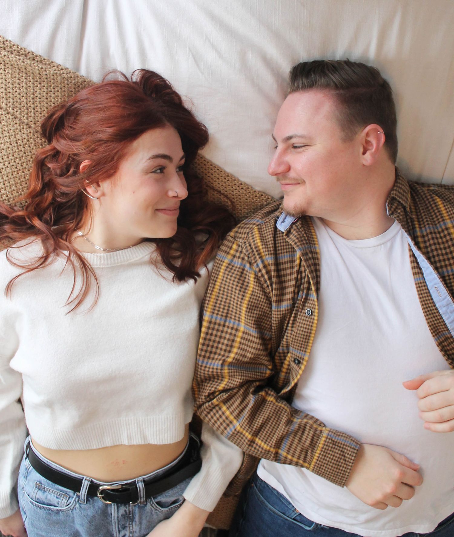 Couple in casual wear lying together on white bedding sharing intimate moments.