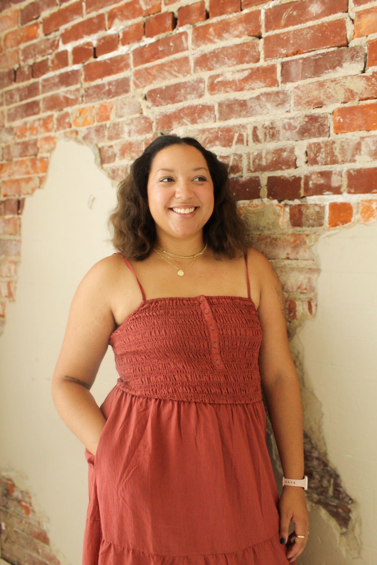A smiling person in a coral-colored sundress poses against a rustic brick wall background.