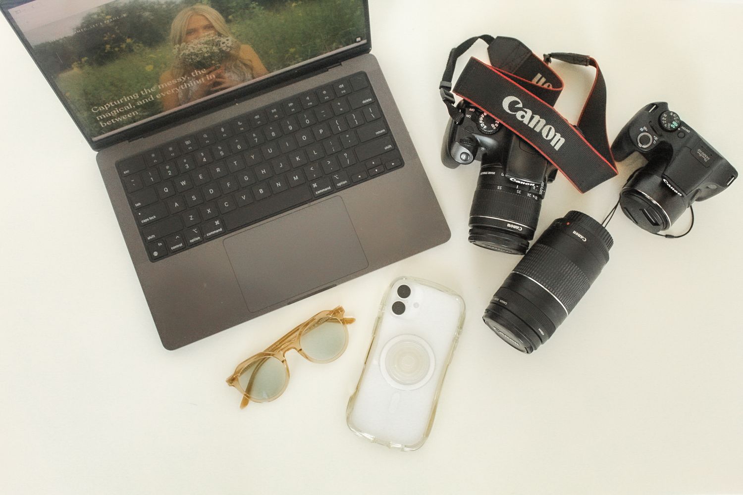 Flatlay of creative workspace with laptop, Canon camera, lenses, iPhone and gold-rimmed glasses on white surface.