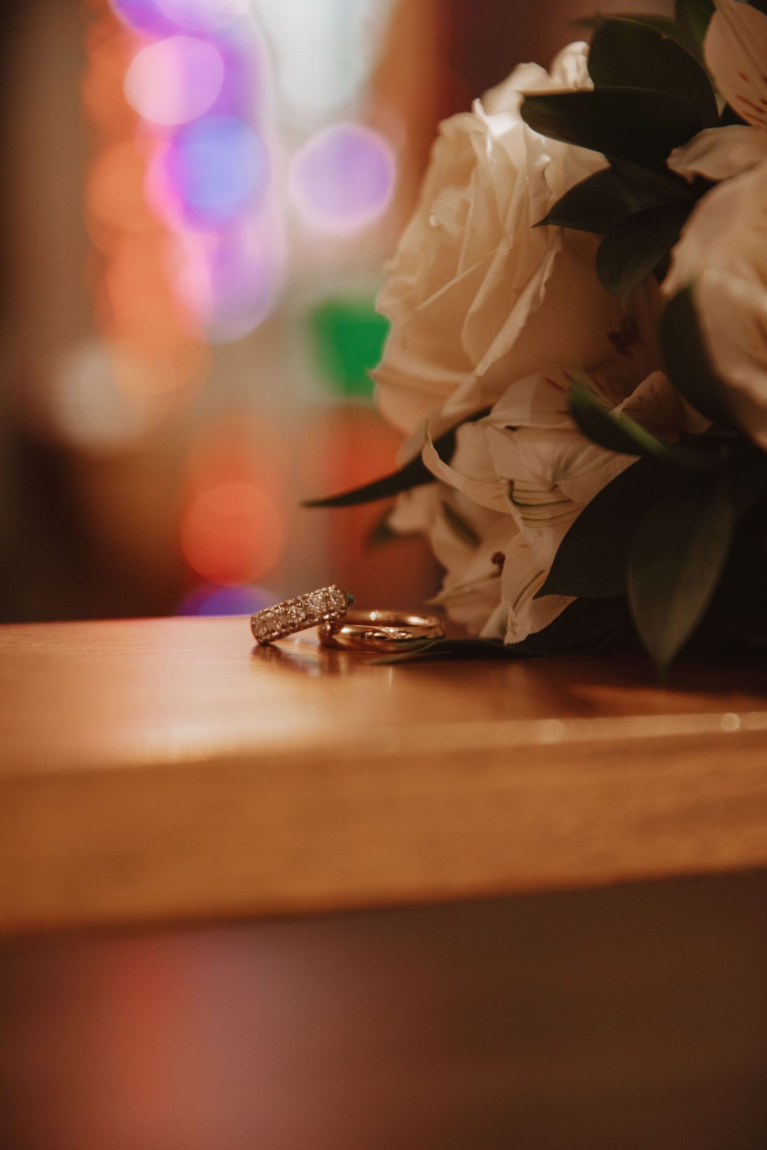 Close-up of white roses and wedding rings with colorful bokeh lighting effects.