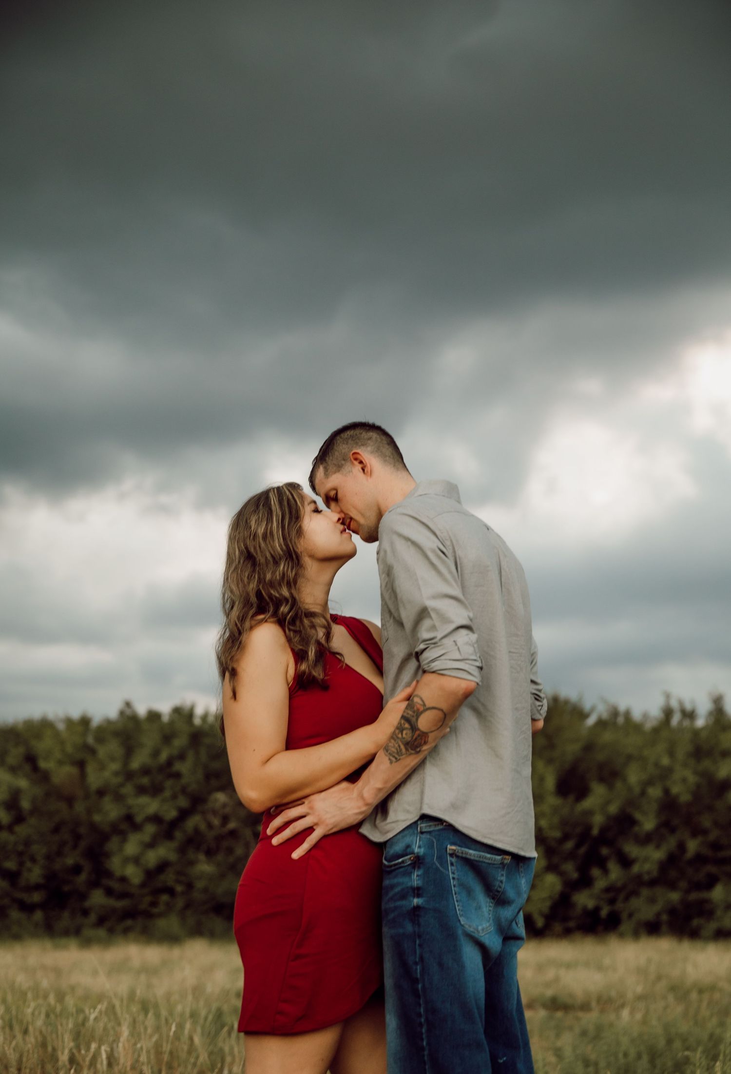A couple shares an intimate moment in a field under dramatic storm clouds.