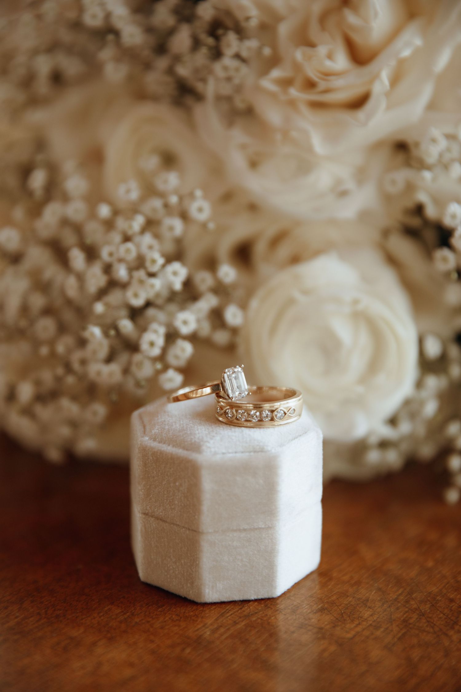 Wedding rings displayed in a white hexagonal box with delicate floral arrangements in the background.
