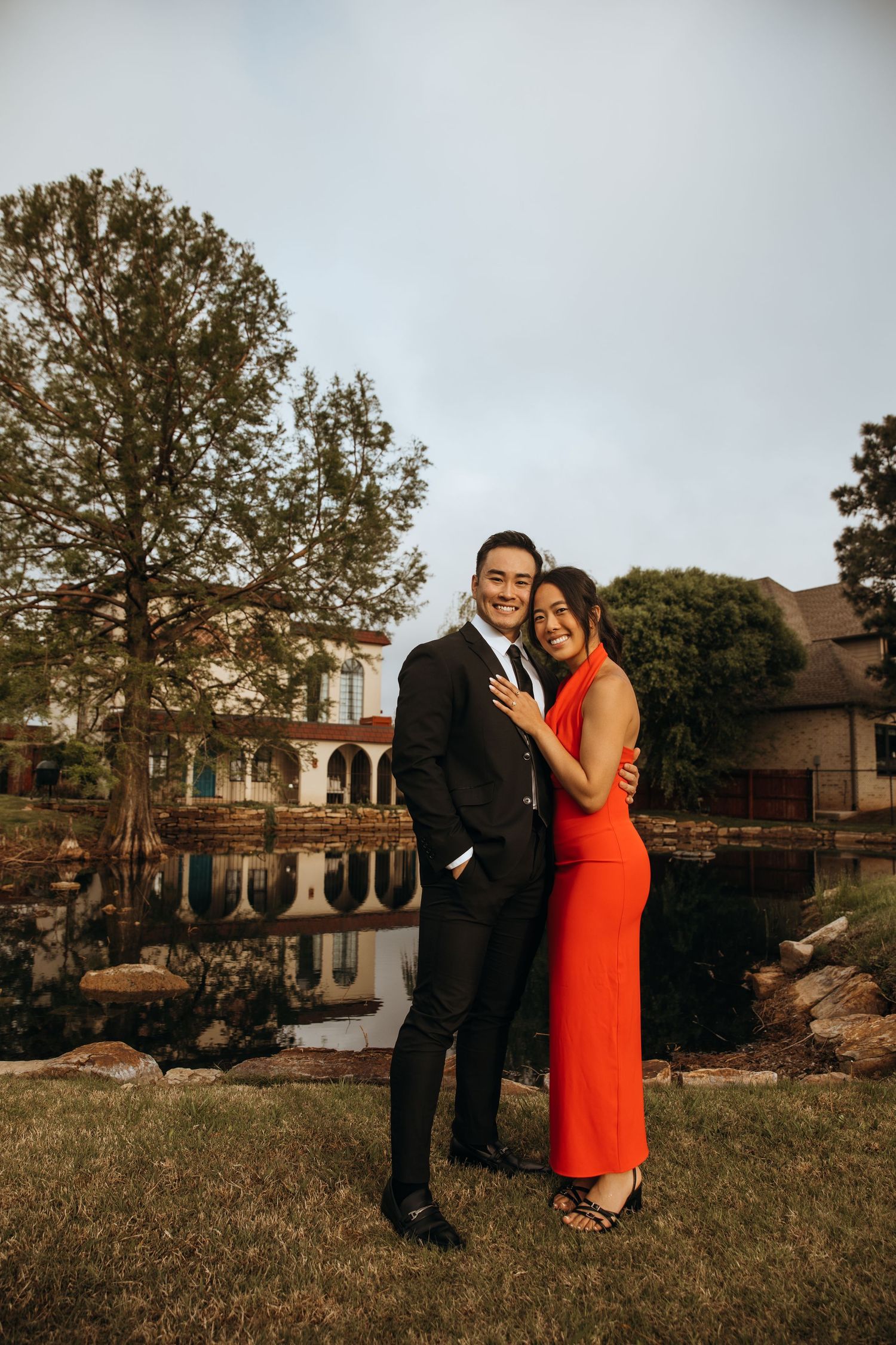 A couple poses for engagement photos on a grassy lawn with houses in the background.