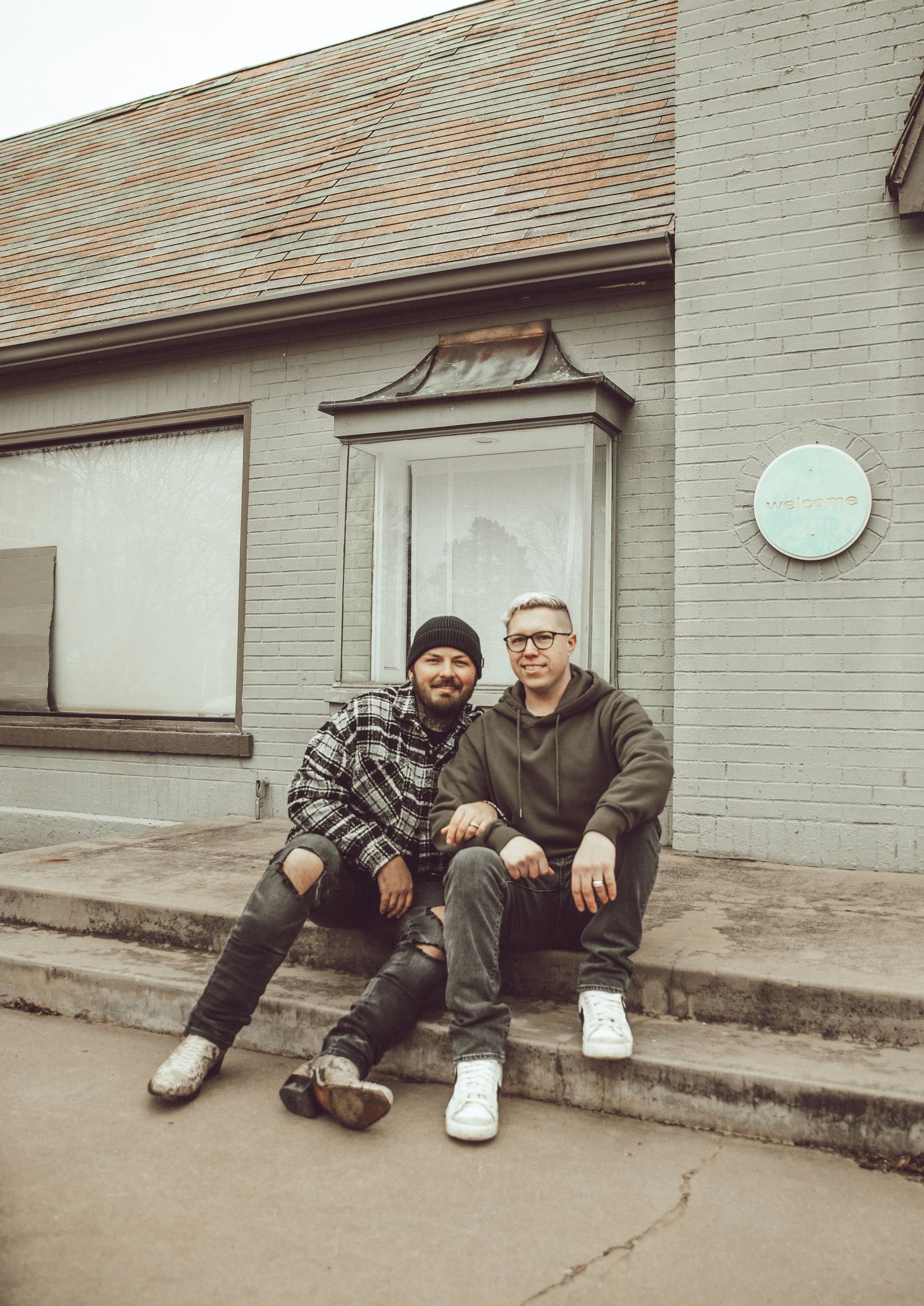 Two people sit casually on concrete steps outside a building with a vintage aesthetic.