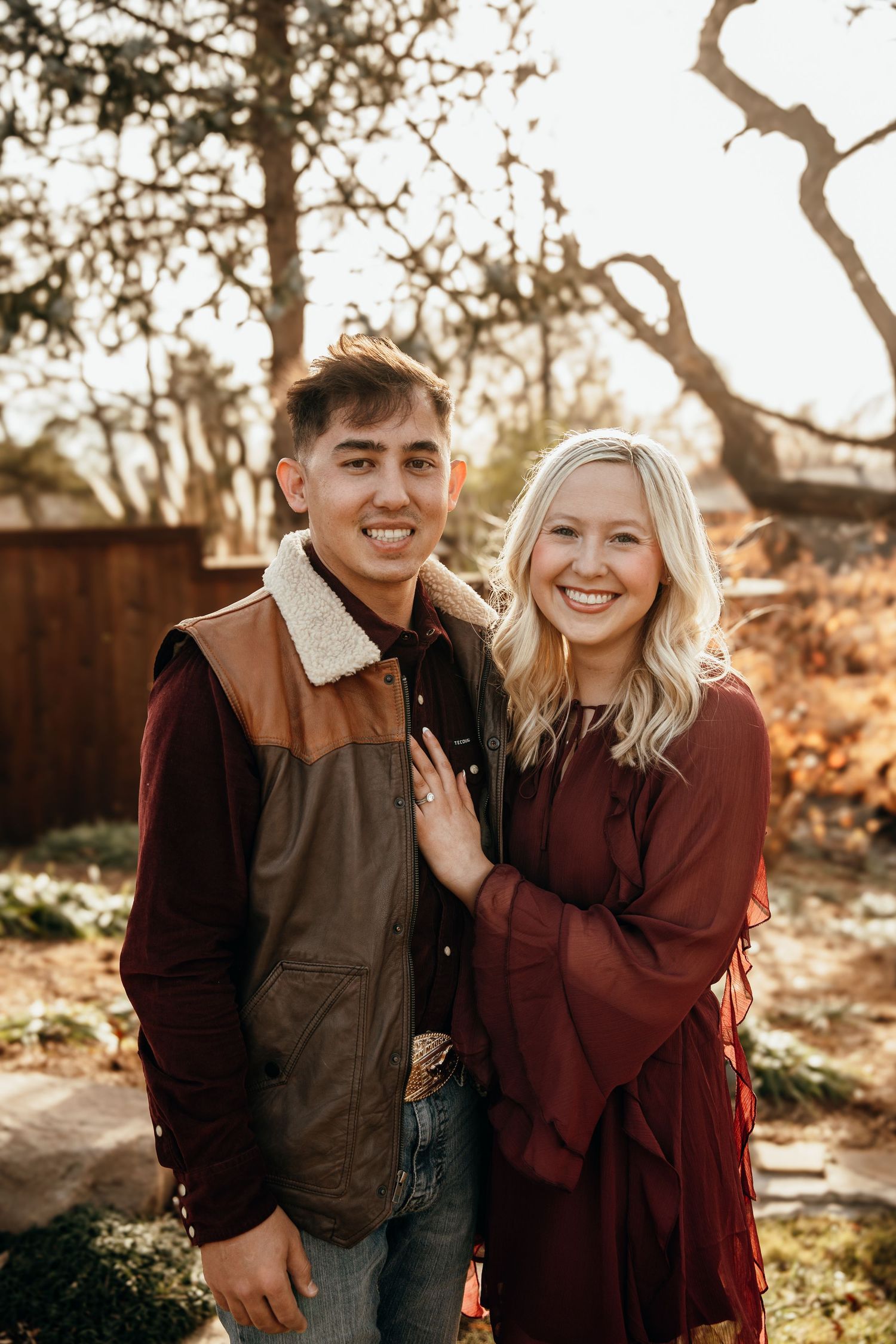 A smiling young couple poses outdoors in burgundy outfits during golden hour with trees in the background.
