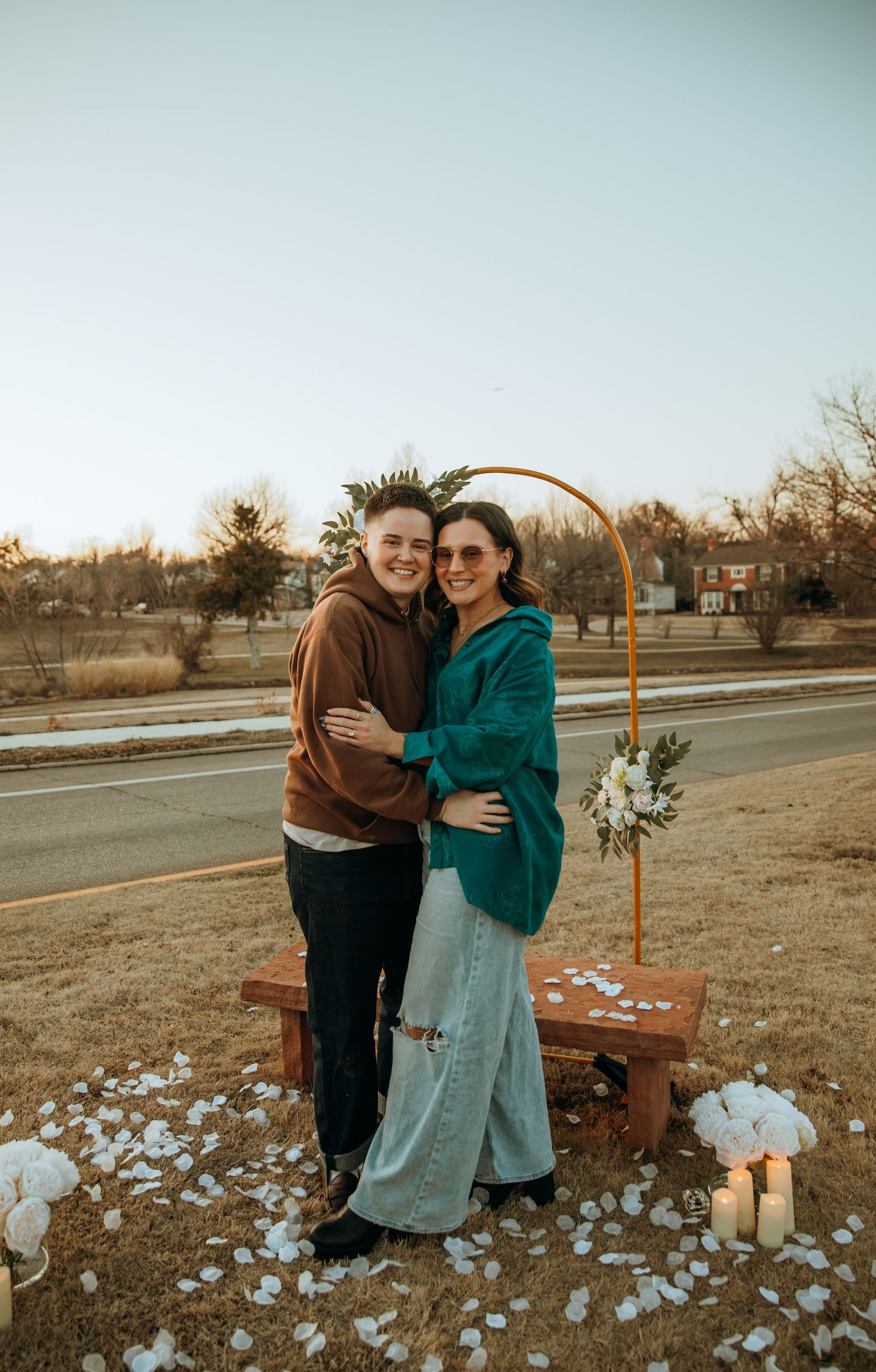 A couple embraces near a rustic arch decorated with white flowers at sunset.