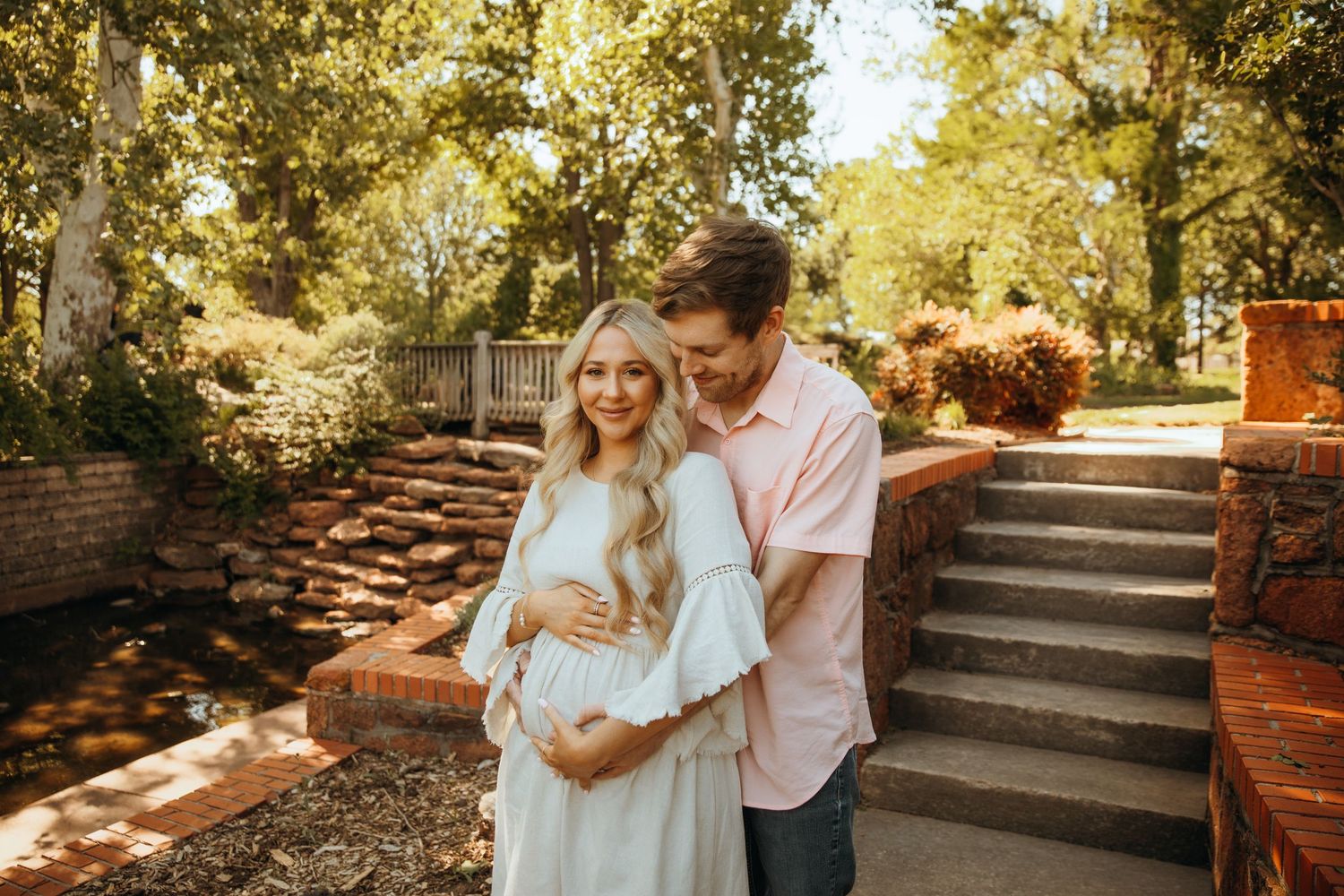 Couple sharing a tender maternity moment outdoors, woman in white dress cradling baby bump, surrounded by lush garden greenery.