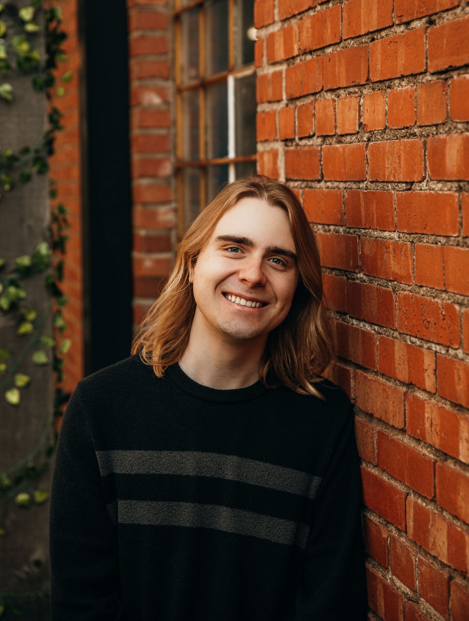 Smiling young man with long blonde hair wearing a black striped sweater, leaning against a red brick wall.