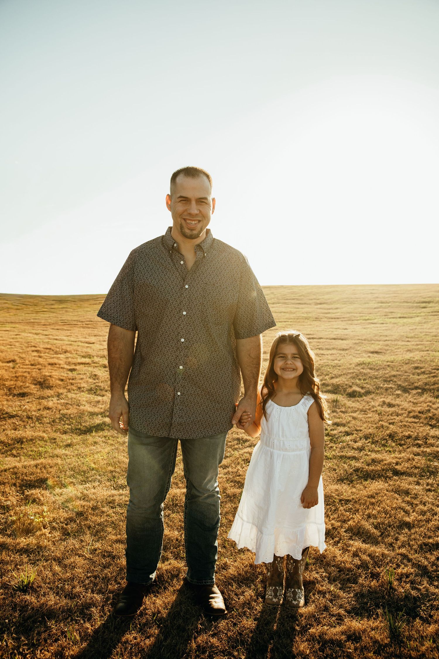 A parent and child hold hands while standing together in a golden sunlit field at sunset.