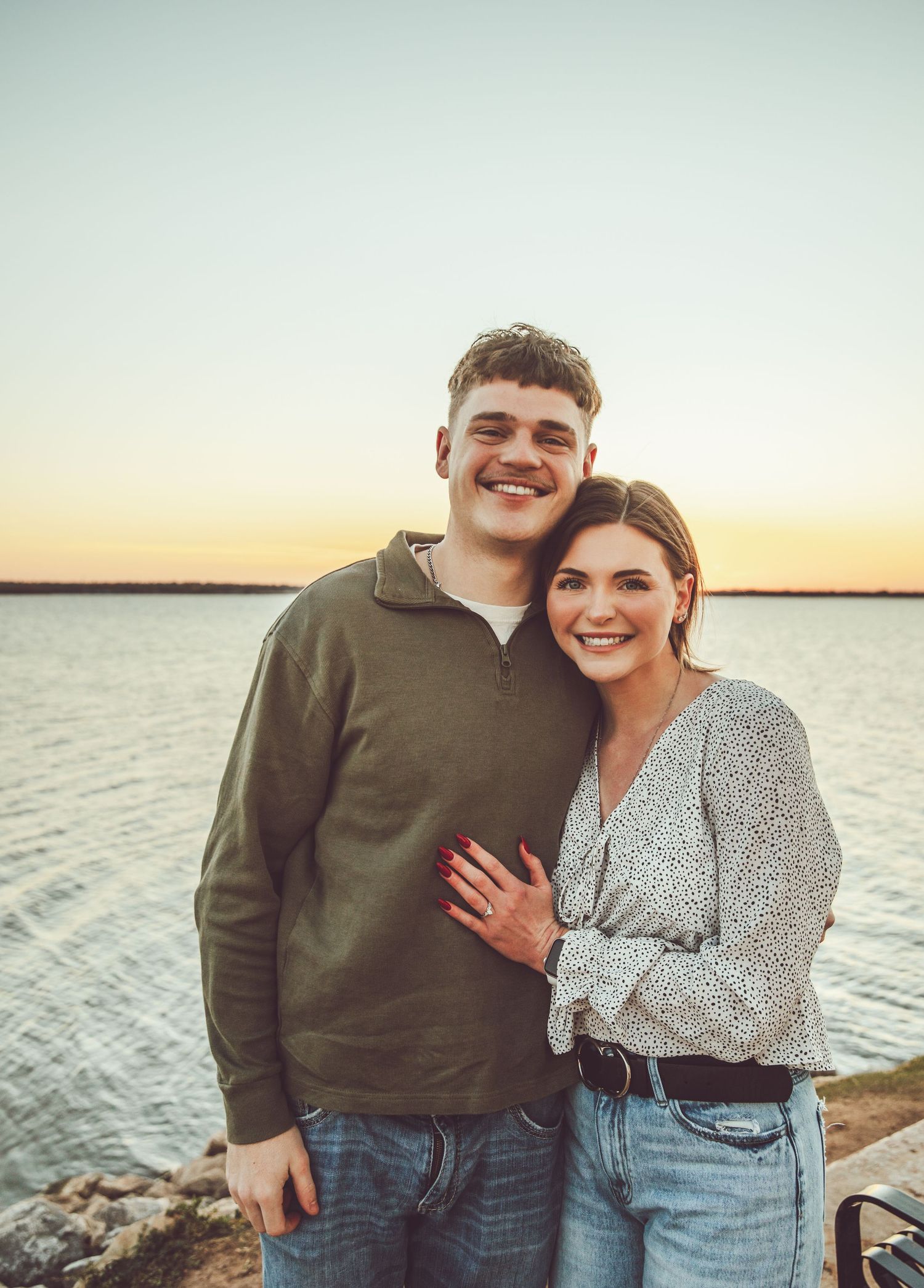 A couple embraces during sunset by the ocean wearing casual denim outfits.