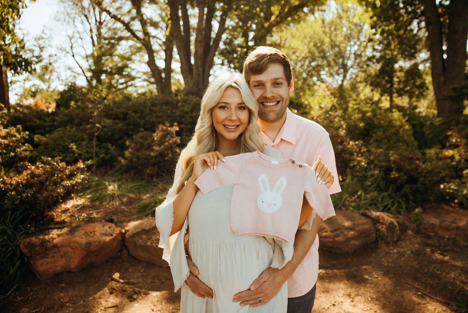 Pregnant couple smiling outdoors holding pink bunny onesie for baby gender reveal in a sunny park setting.