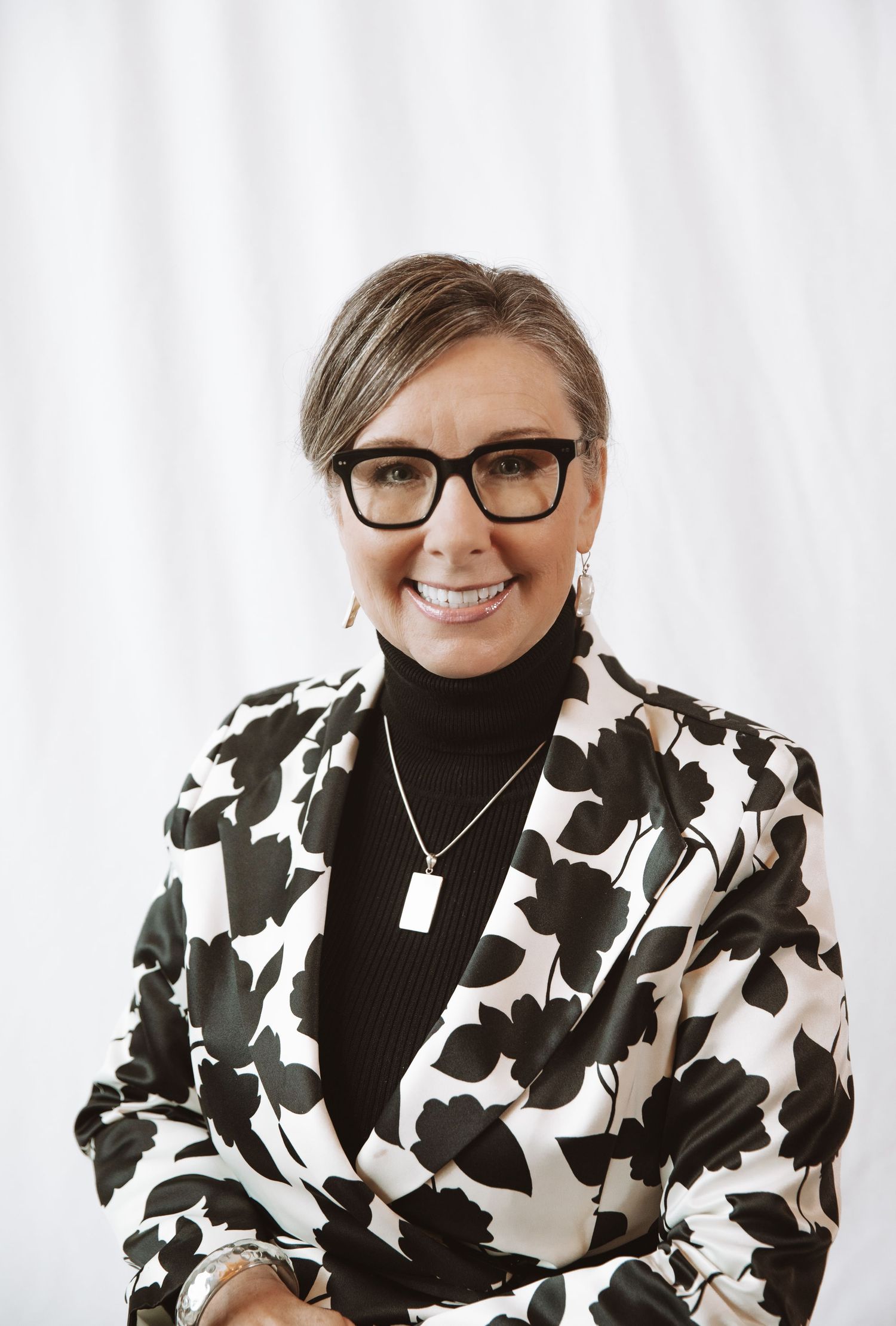 Professional wearing a black and white patterned blazer with black turtleneck and glasses smiles at camera.
