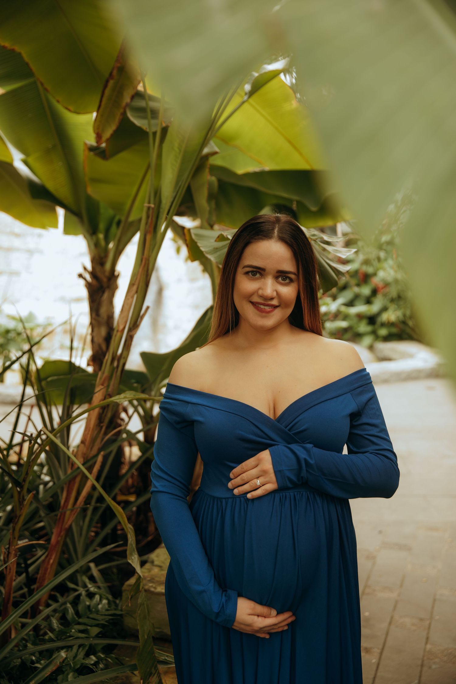 Maternity photo of a woman in a blue off-shoulder dress standing by tropical plants in natural lighting.
