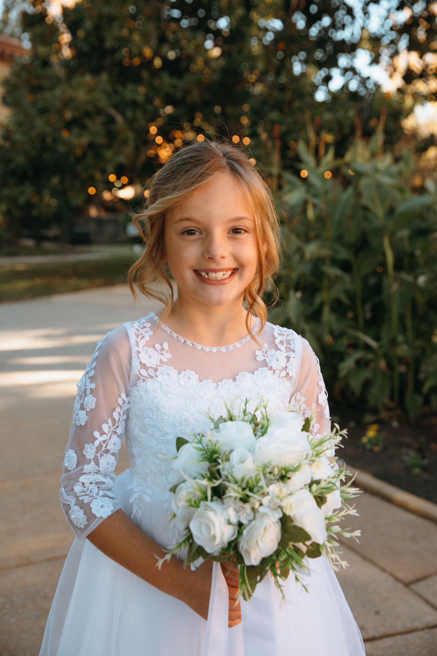 Young flower girl in white lace dress holds elegant white floral bouquet.