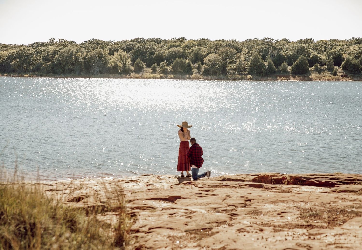 A romantic proposal scene on a rocky lakeside overlook with sparkling water and trees in the background.