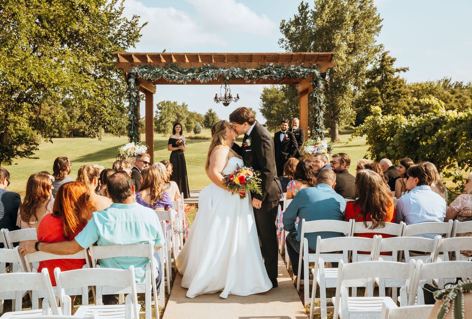 Bride and groom kiss at outdoor wedding ceremony under floral pergola with seated guests on white chairs.