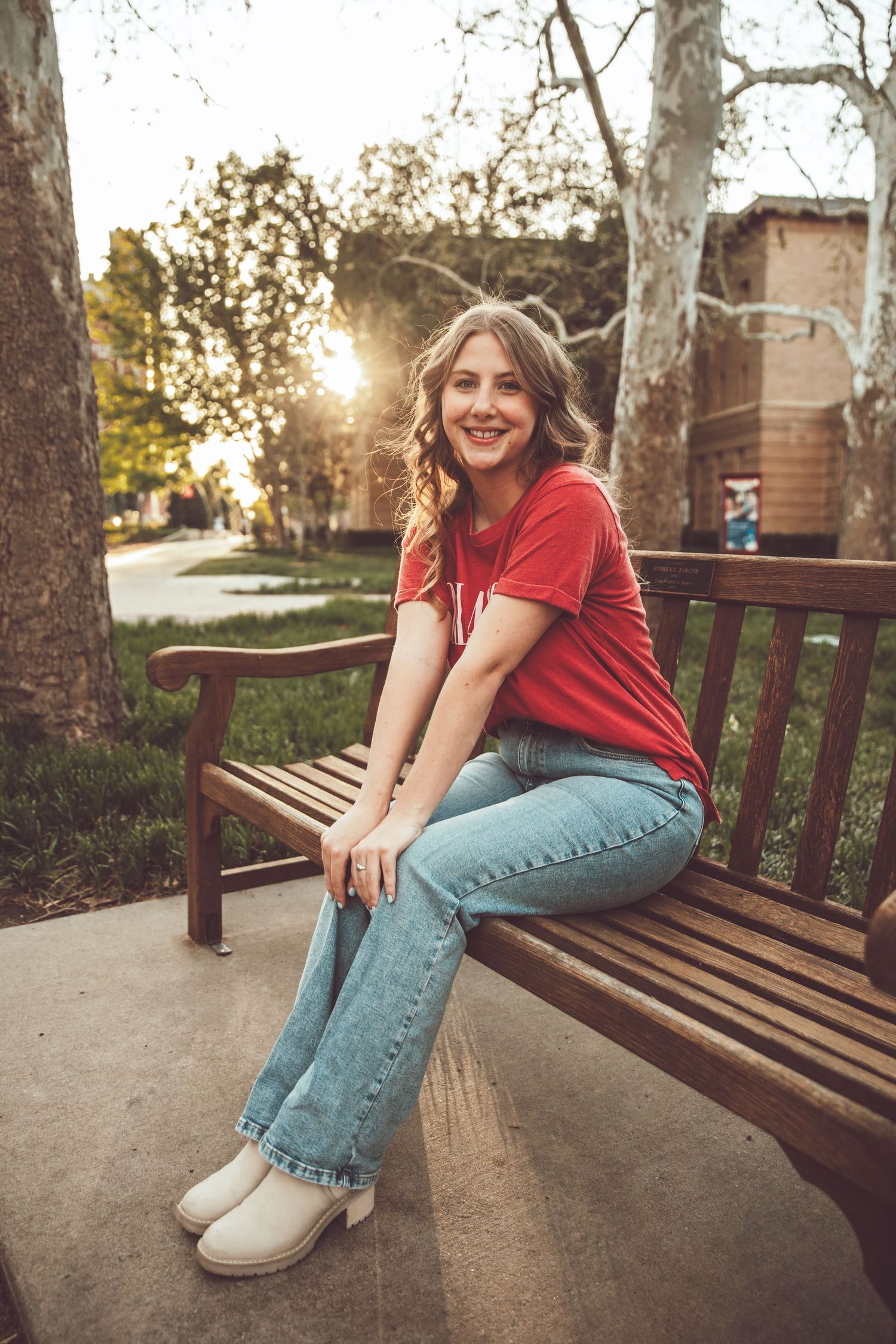 Young woman in red t-shirt and jeans sits smiling on a wooden park bench at golden hour sunset.