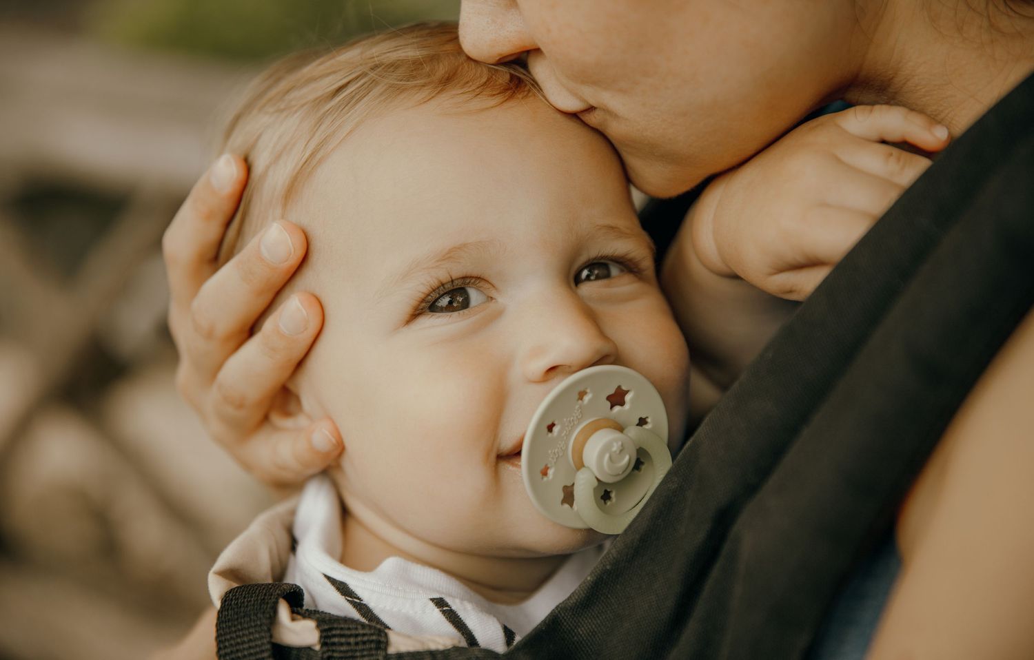 A mother tenderly kisses her baby with a pacifier, cradled in a carrier.
