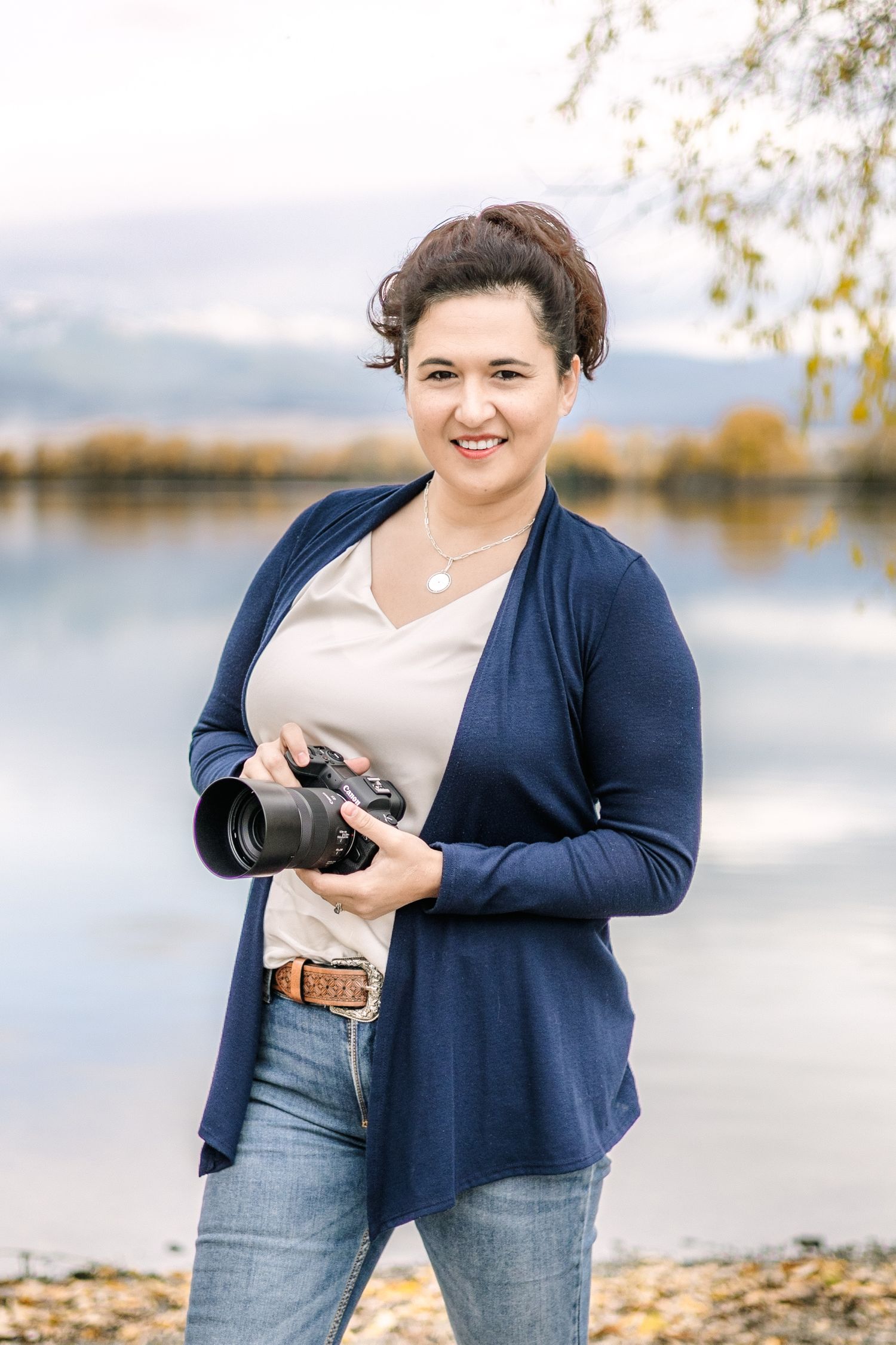 A Helena wedding photographer in a navy cardigan and jeans poses with a camera near a lake on an autumn day.