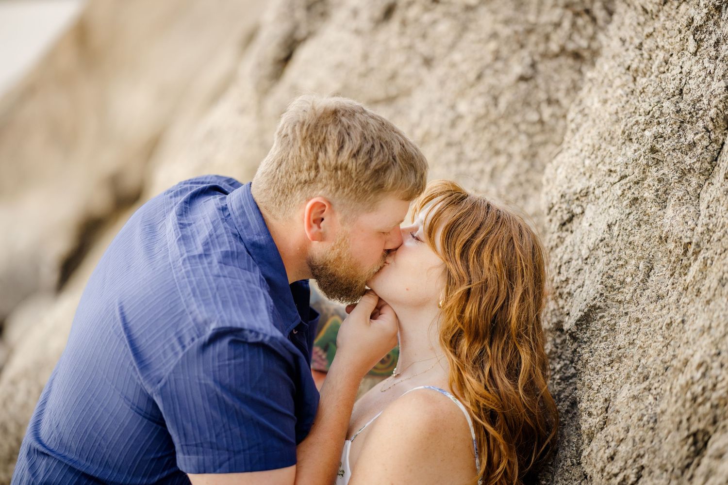 Couple sharing a romantic kiss against a rocky wall outdoors, man in blue shirt with redhead woman.