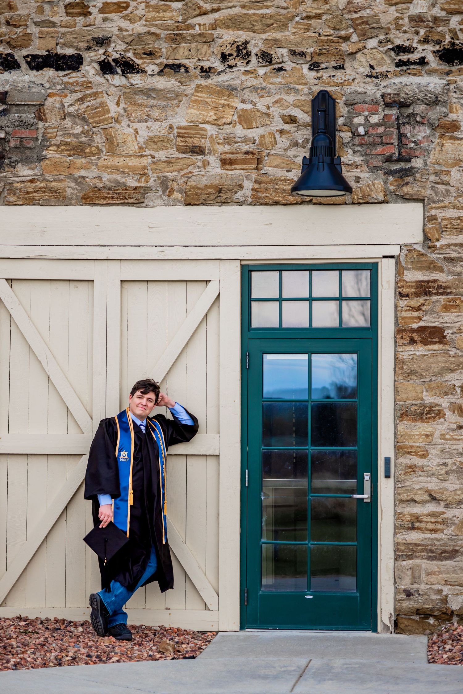 Bozeman Senior in cap and gown by a stone building exterior with glass door and colored barn door. A portrait by Kelsea Laine Photography