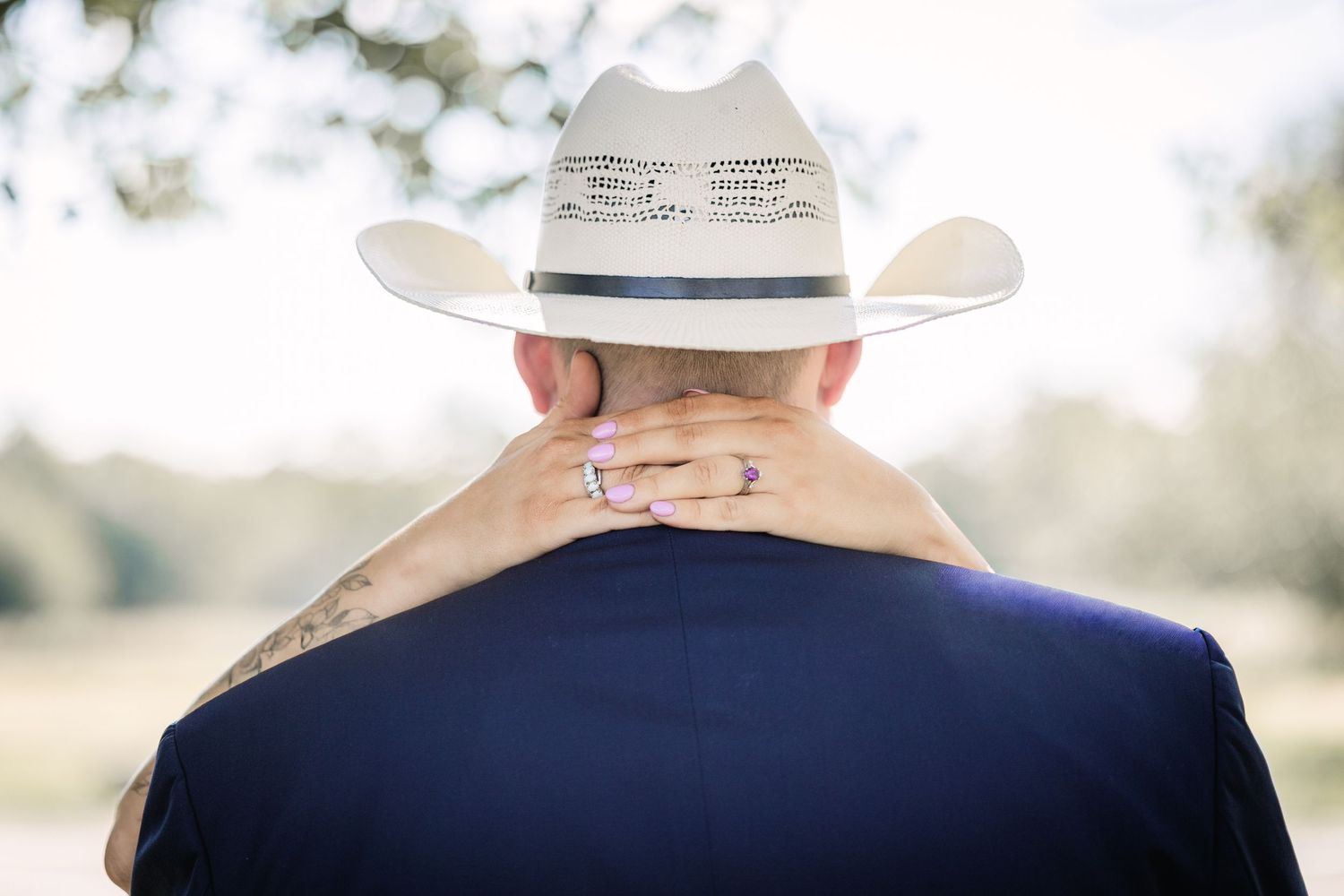 Couple embracing outdoors, man wearing white cowboy hat and blue shirt, woman's hand with purple nails on his neck.